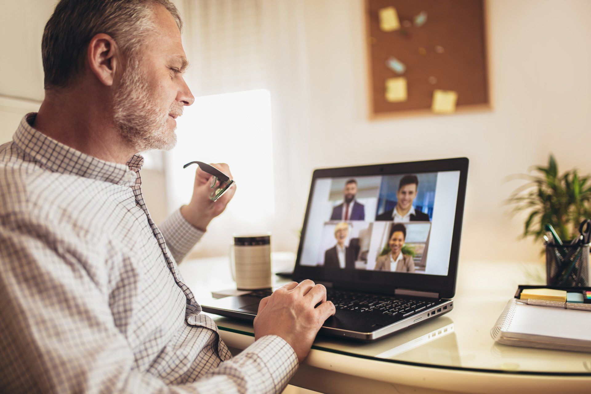 A person in a plaid shirt sits at a desk, looking at a laptop displaying a video conference call with four colleagues.