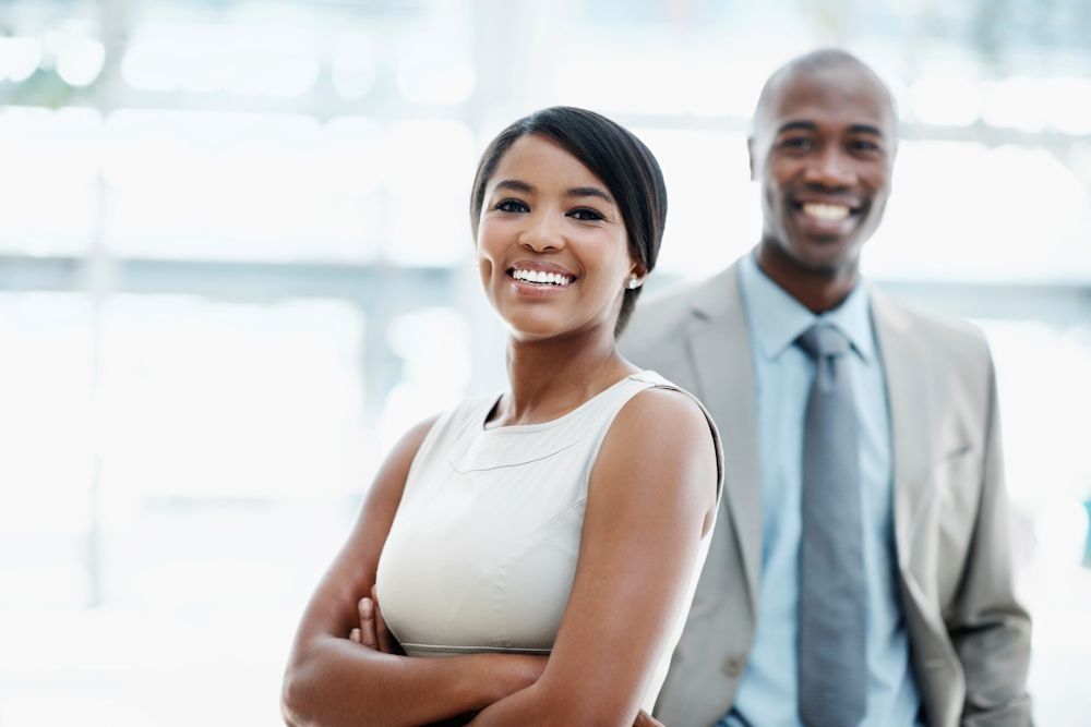 A smiling professional standing with arms crossed in front of a colleague in a suit, set in a bright office environment.
