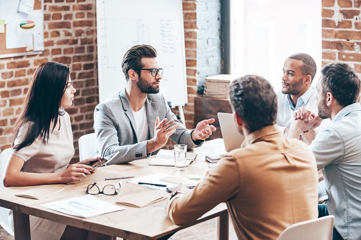 Five professionals collaborate in an office meeting around a wooden table, engaged in active discussion.
