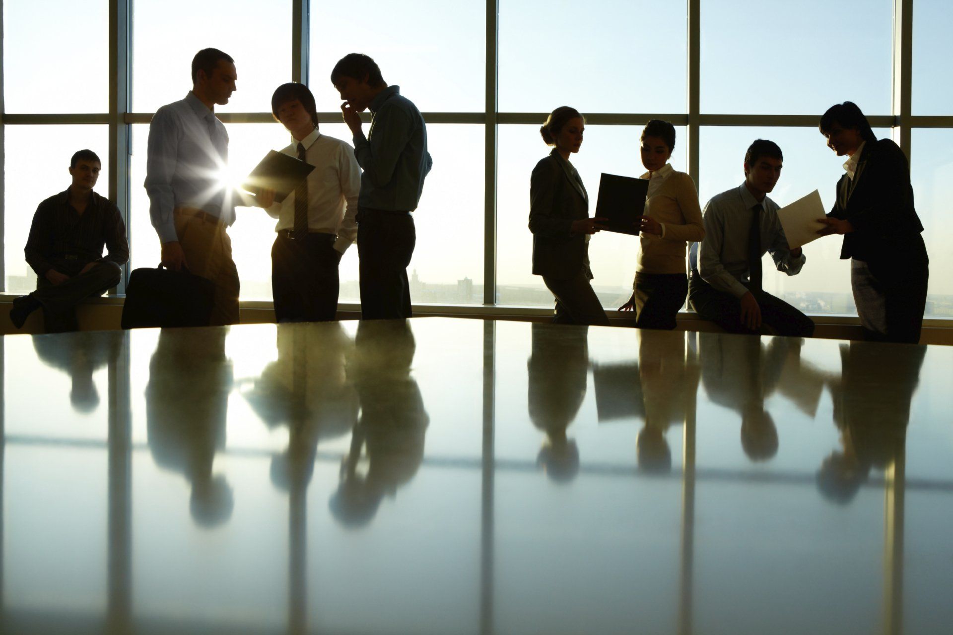 Silhouetted people standing in a sunlit office meeting, reflecting on a polished table.
