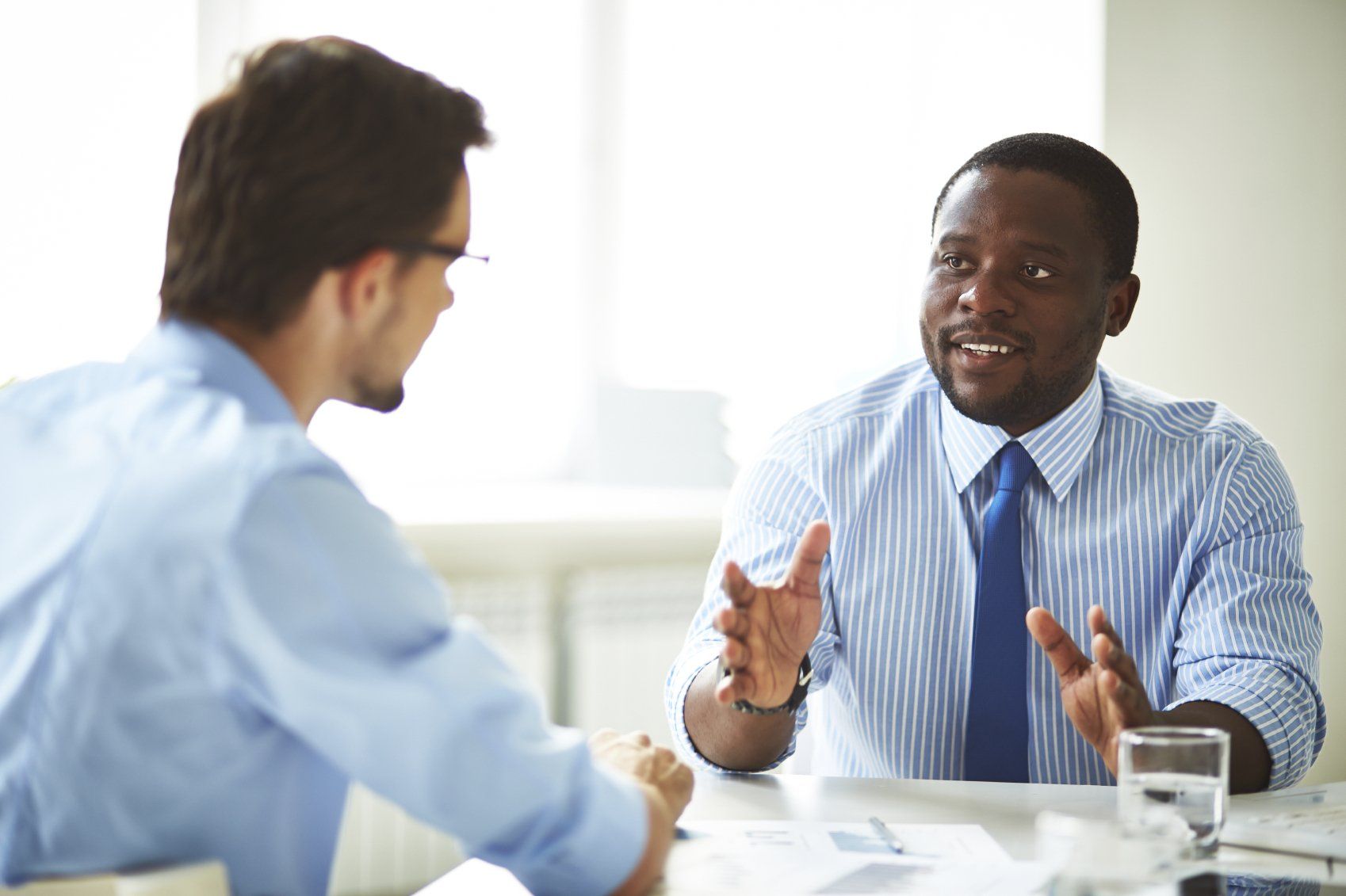 Two professionals in button-down shirts discuss business at a bright office table, one gesturing during conversation.