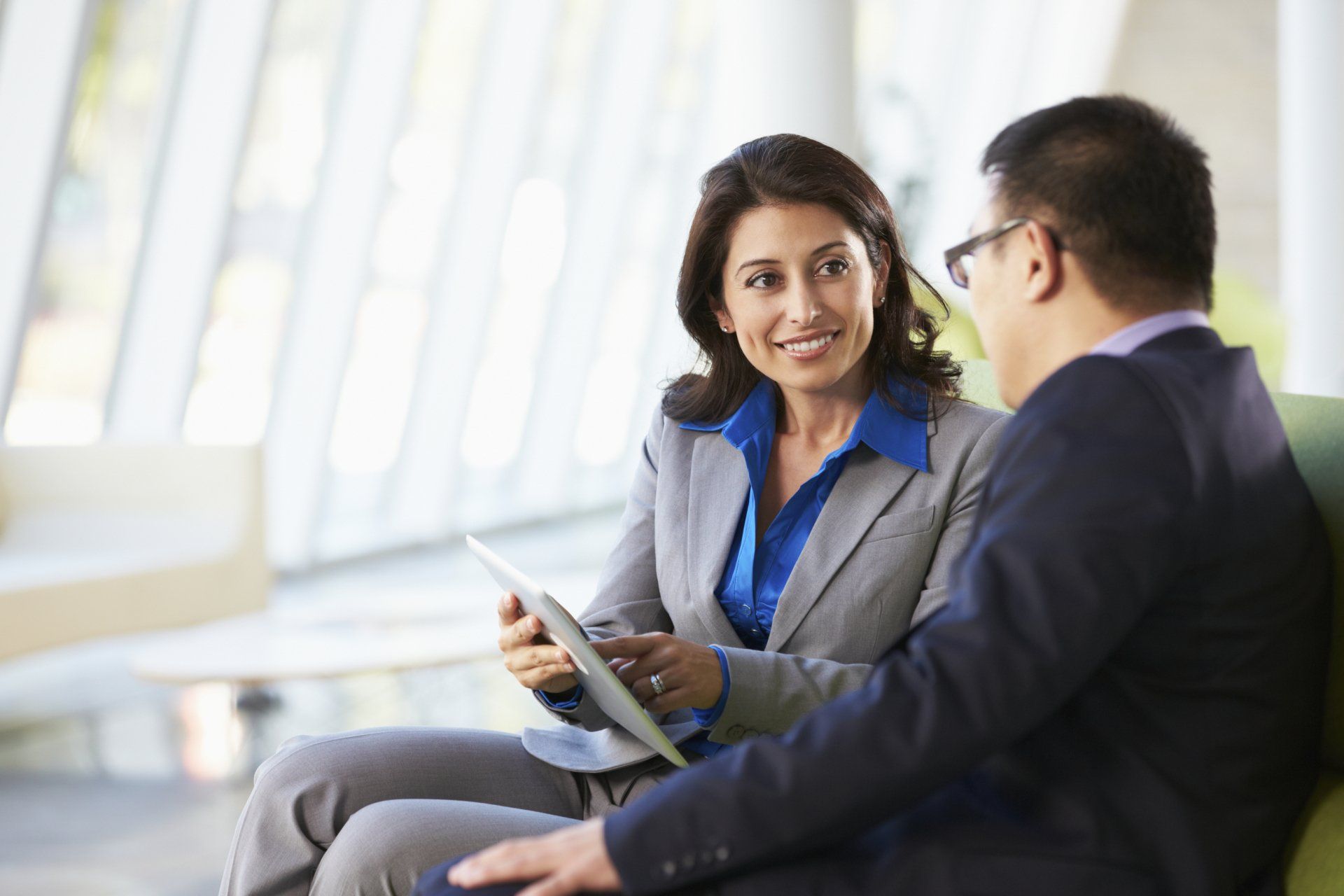 A person in a gray blazer shows a tablet to a colleague in a navy suit while sitting in a bright, modern office lobby.