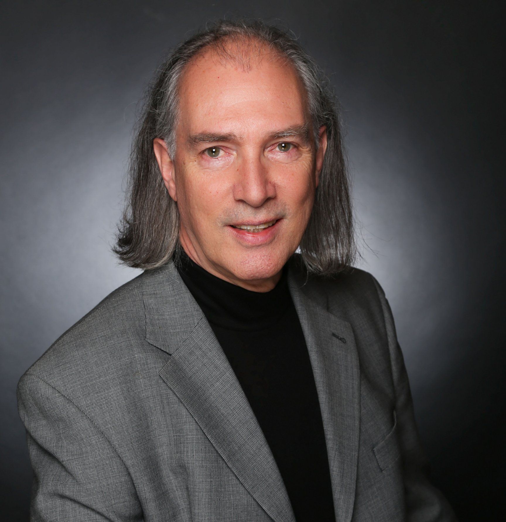 A person with shoulder-length grey hair, wearing a black turtleneck and grey patterned blazer against a dark background.