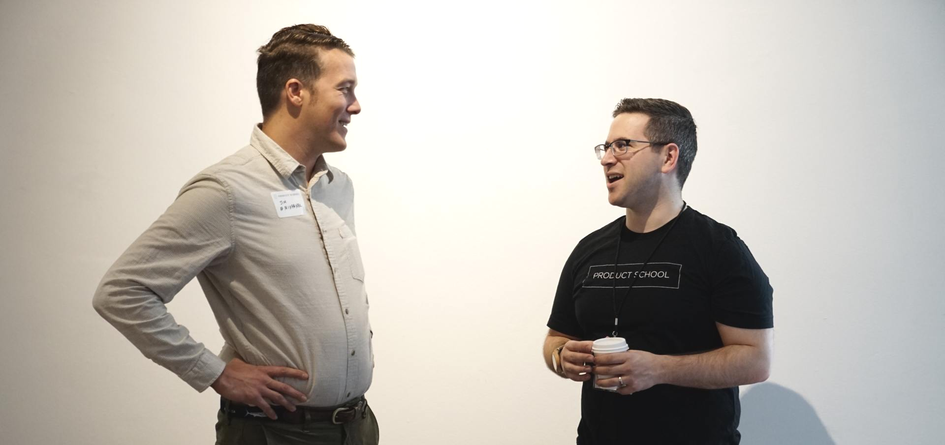 Two people standing against a plain white wall, conversing while looking at each other.