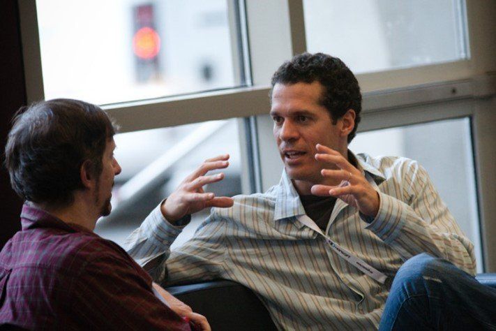 Two people sit on a couch in front of a window, one gesturing with both hands while speaking to the other.
