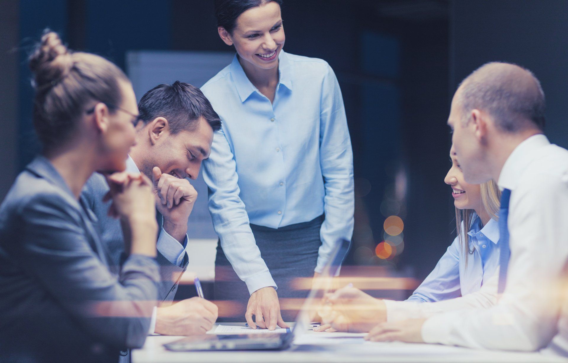 A group of professionals in business attire collaborate around a desk while reviewing documents in an office setting.