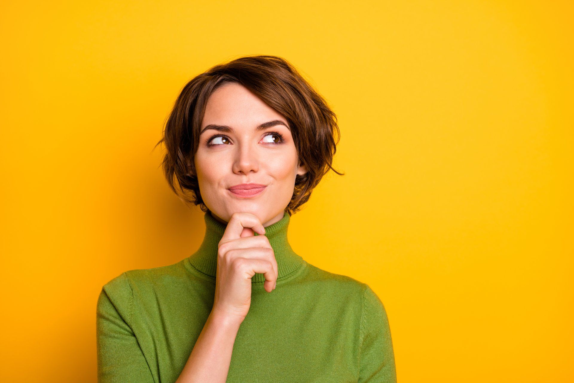 A person in a green turtleneck sweater looking up thoughtfully with a hand on their chin against a bright yellow background.