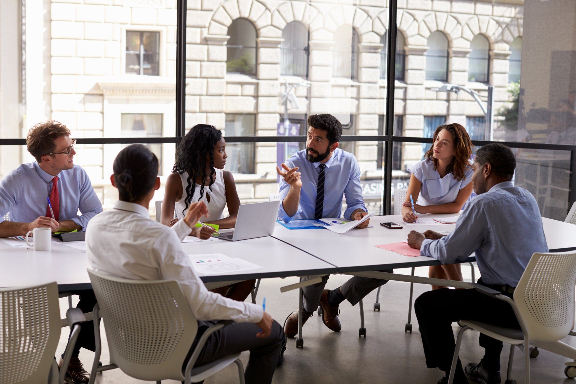 A diverse group of professionals in business attire hold a collaborative meeting around a conference table in an office.