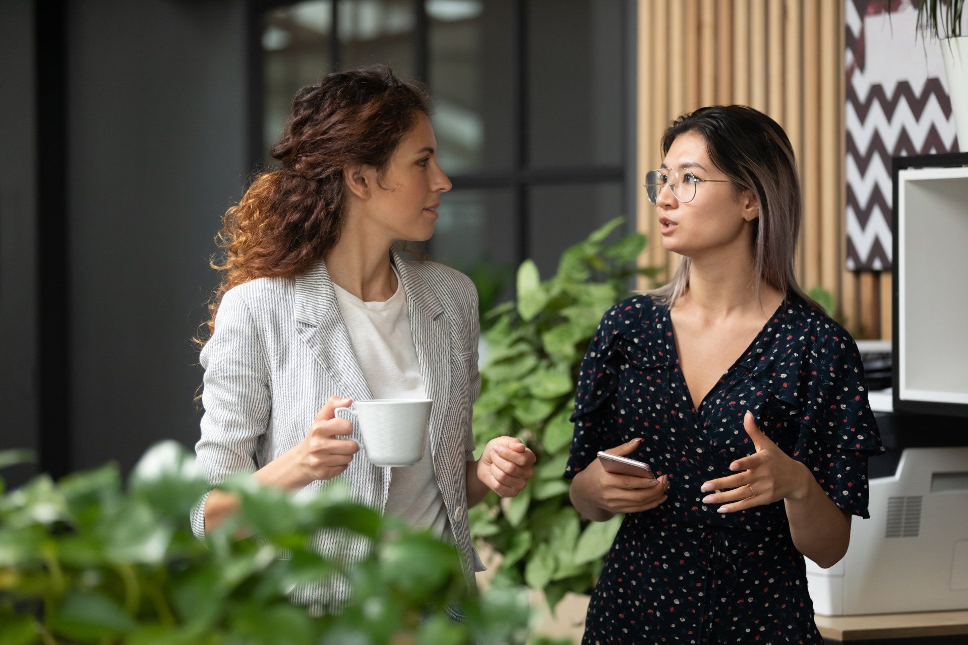 Two colleagues talk in an office setting, one holding a mug and the other gesturing while holding a smartphone.