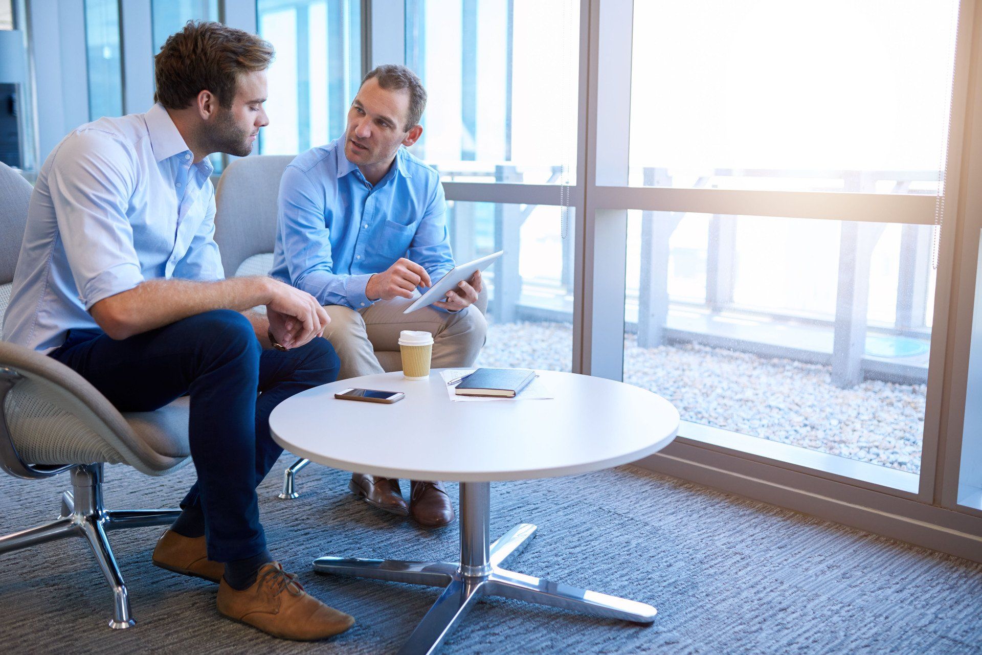 Two professionals in button-down shirts discuss data on a tablet while sitting in a bright, modern office lounge area.