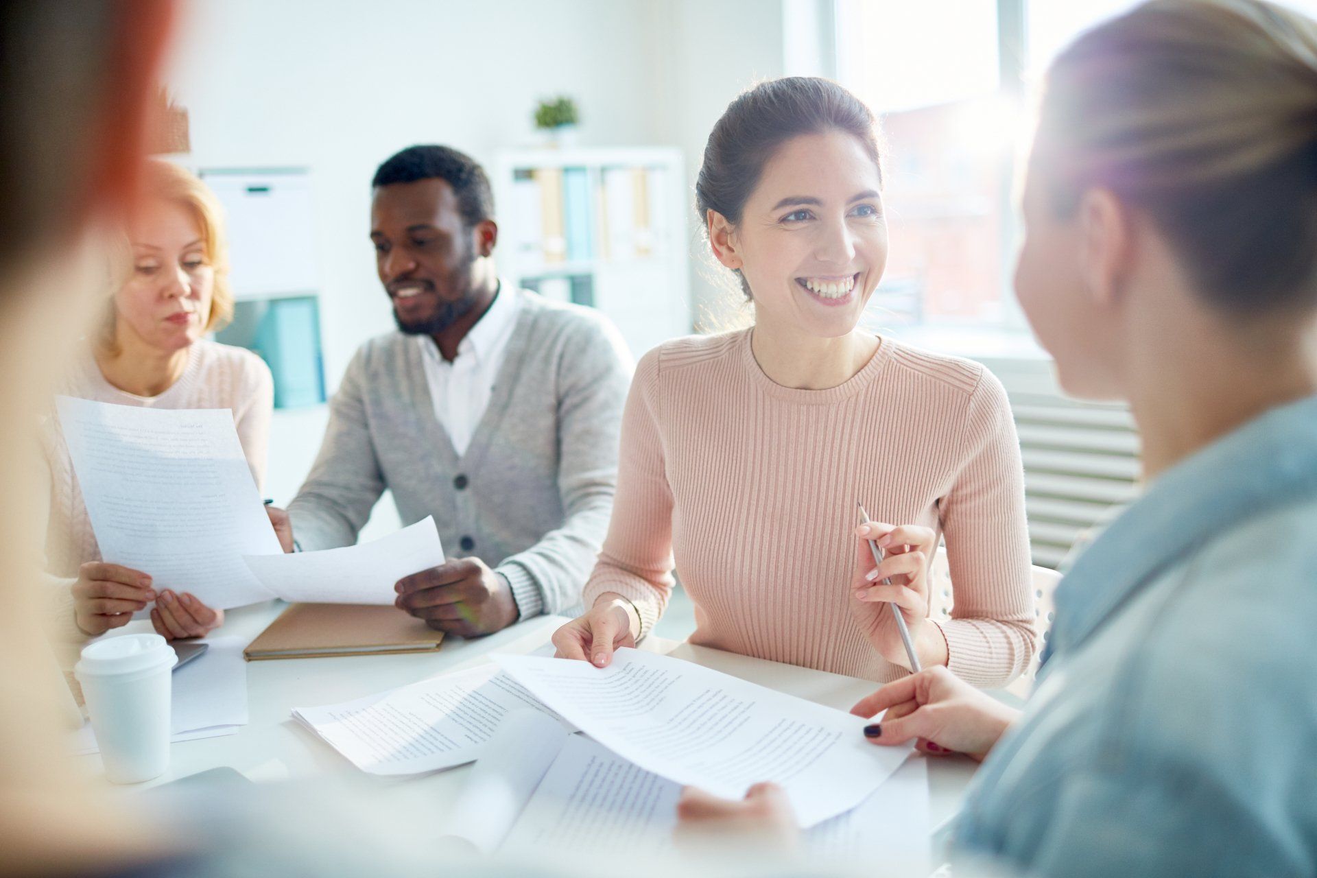 A diverse group of colleagues sit around a table in a bright office, reviewing documents and talking with smiles.
