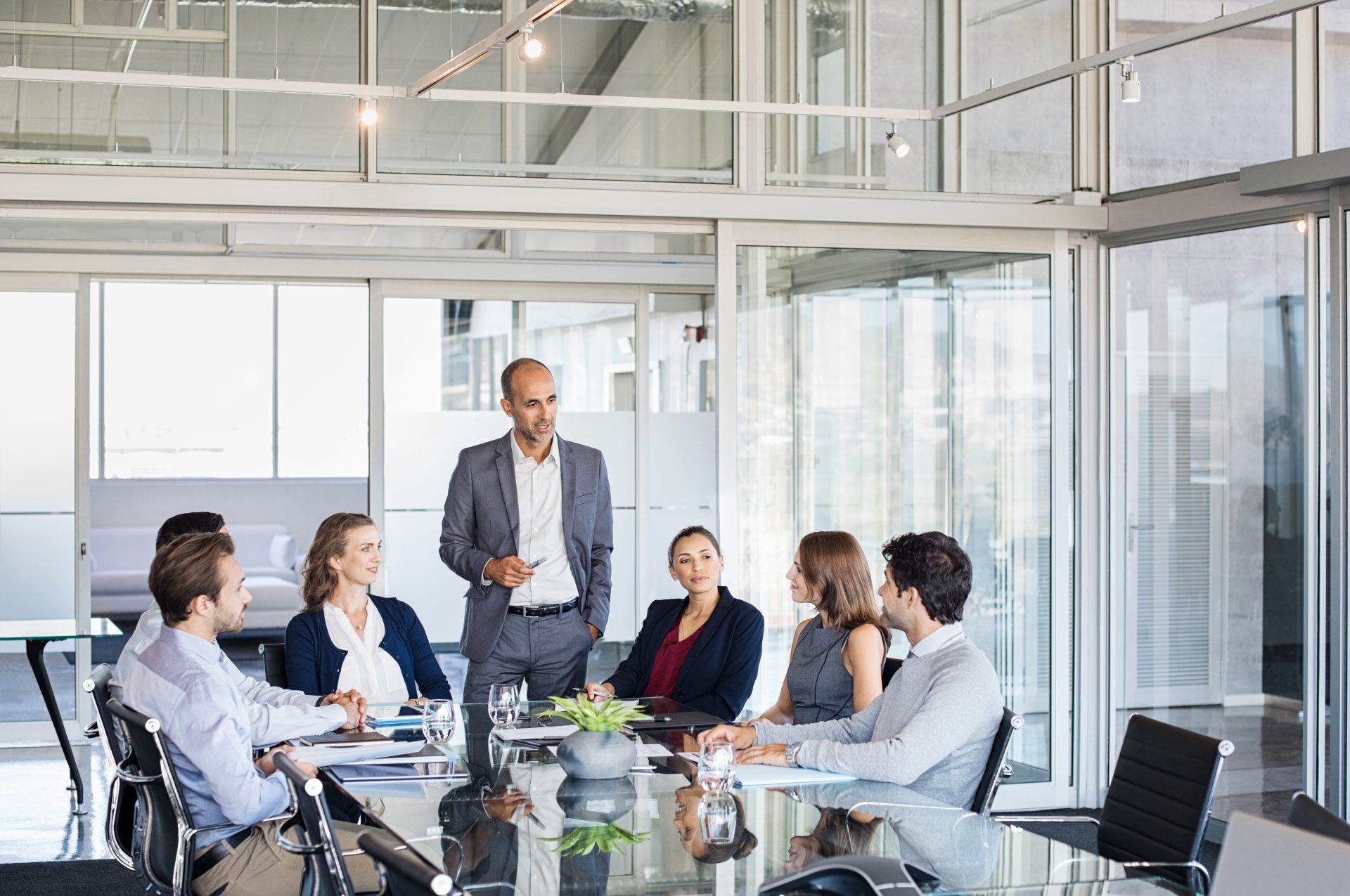 A group of professionals in business attire meet in a brightly lit, glass-walled conference room.