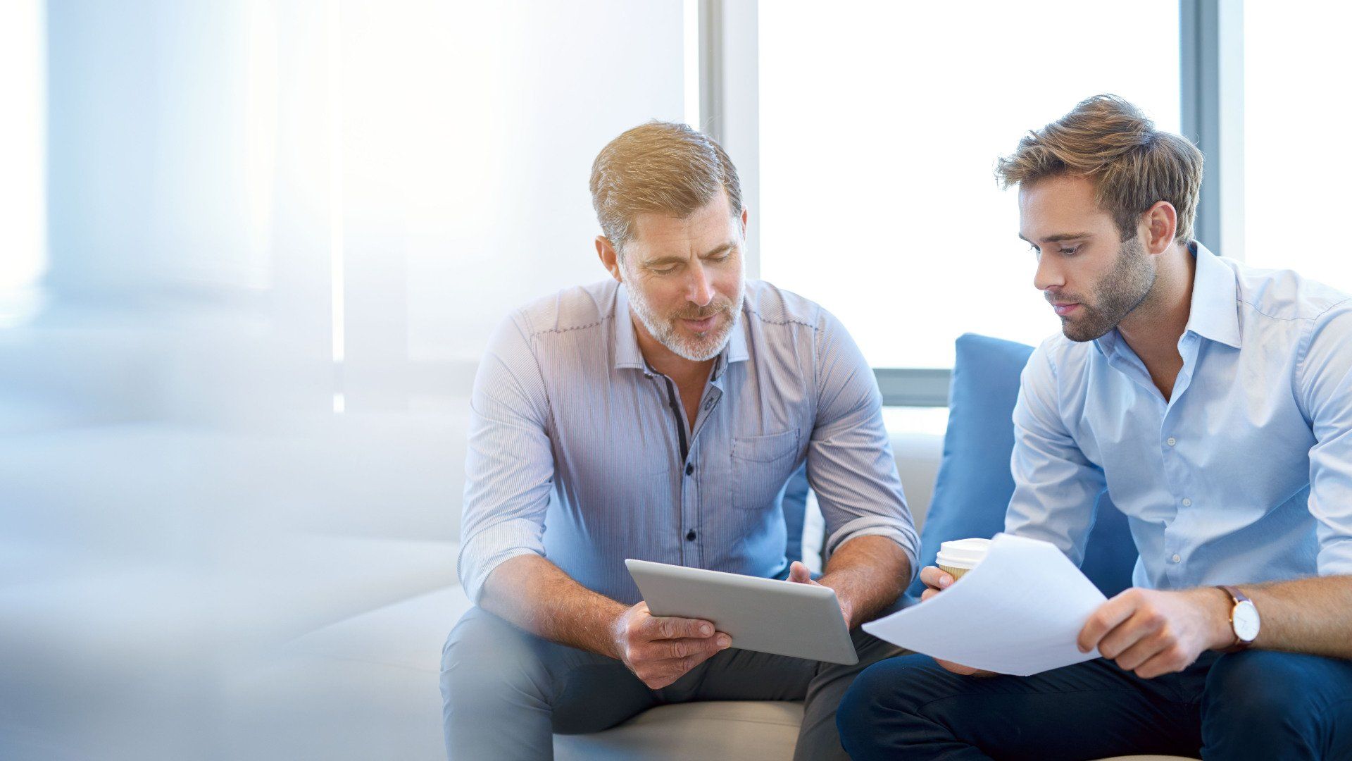 Two men in light blue shirts sit in a brightly lit office, reviewing a tablet and documents together.