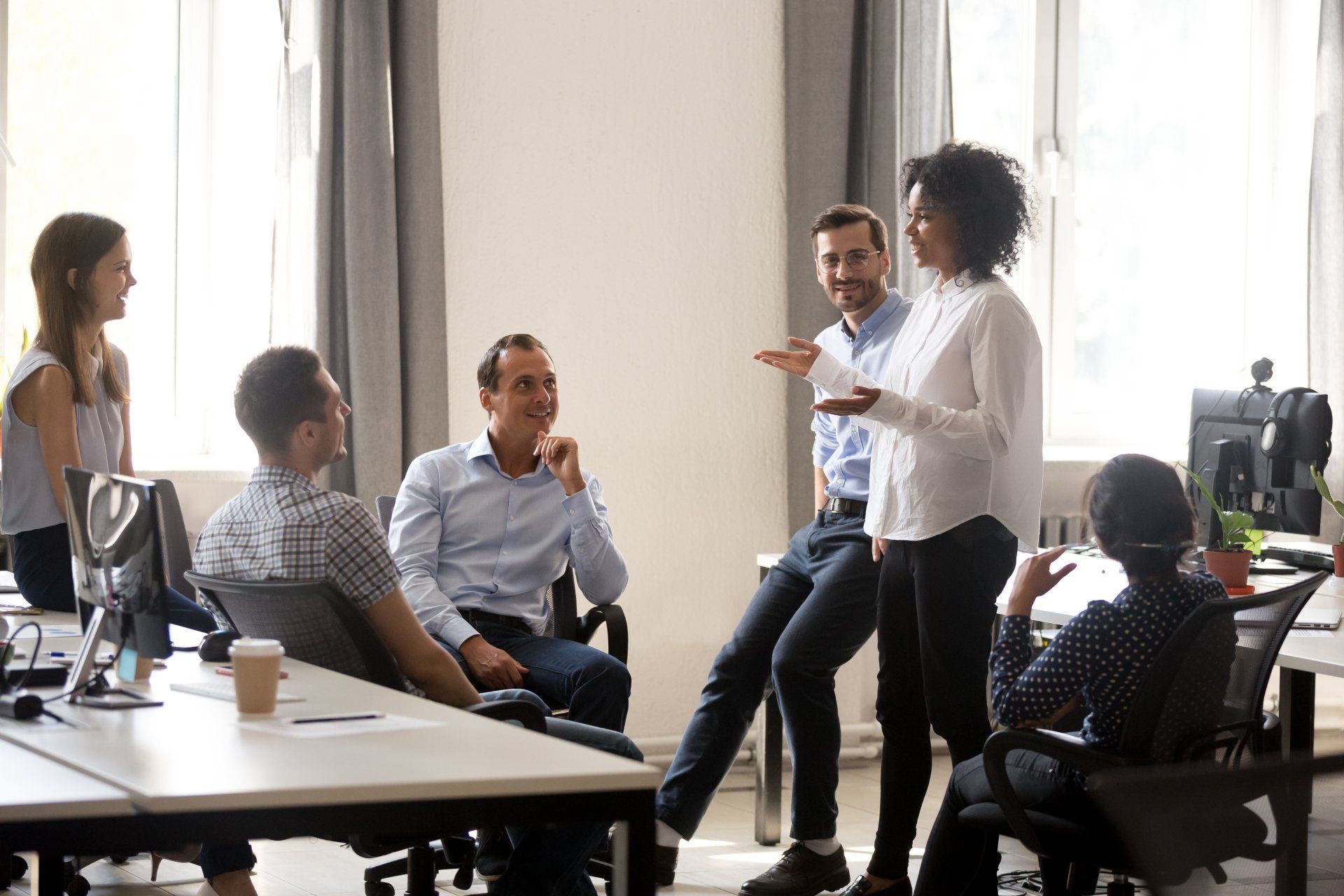 A diverse group of colleagues gather in a bright, modern office to discuss work during a casual meeting.