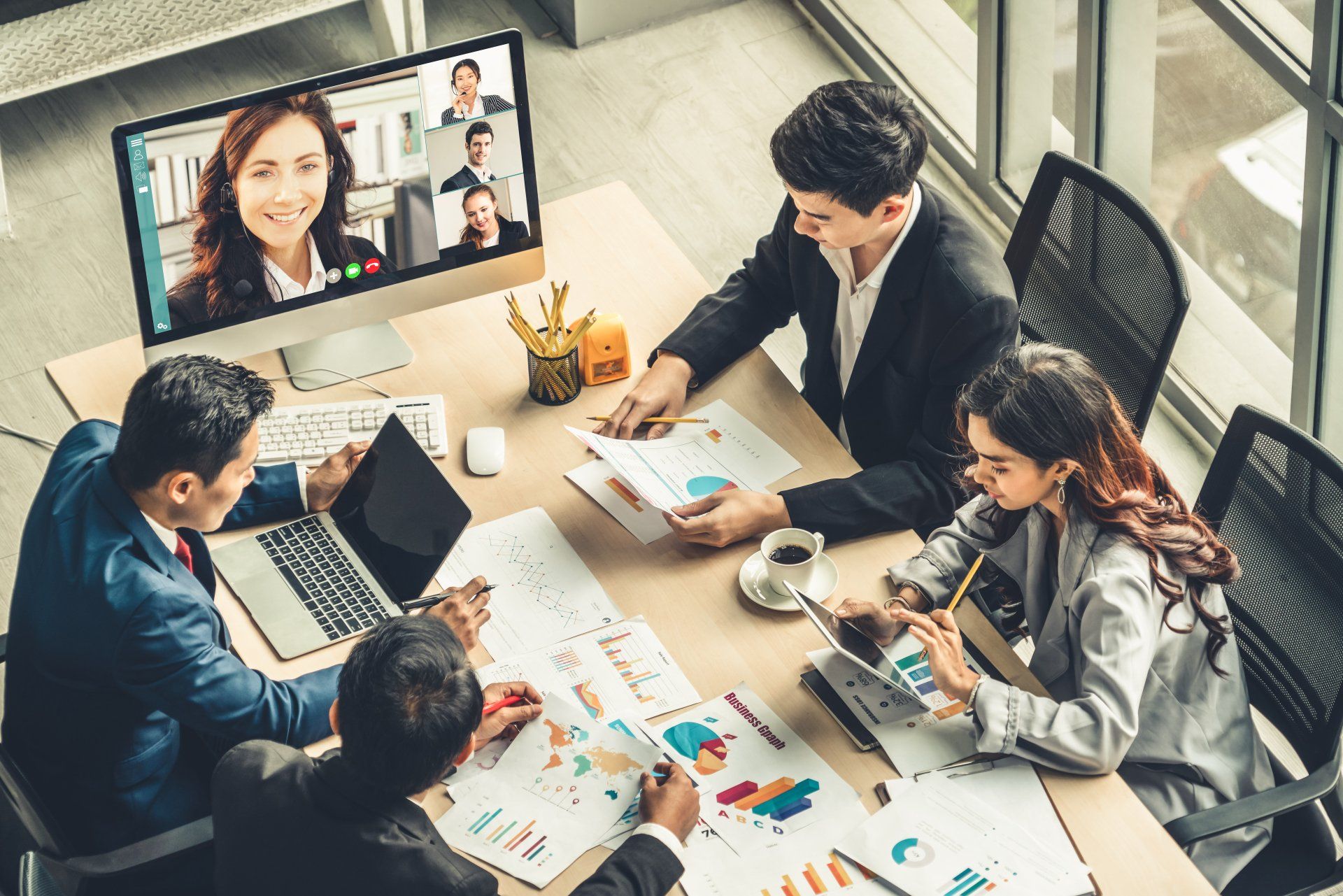 A team meets at a table with papers and a laptop, while viewing remote colleagues on a desktop monitor during a meeting.