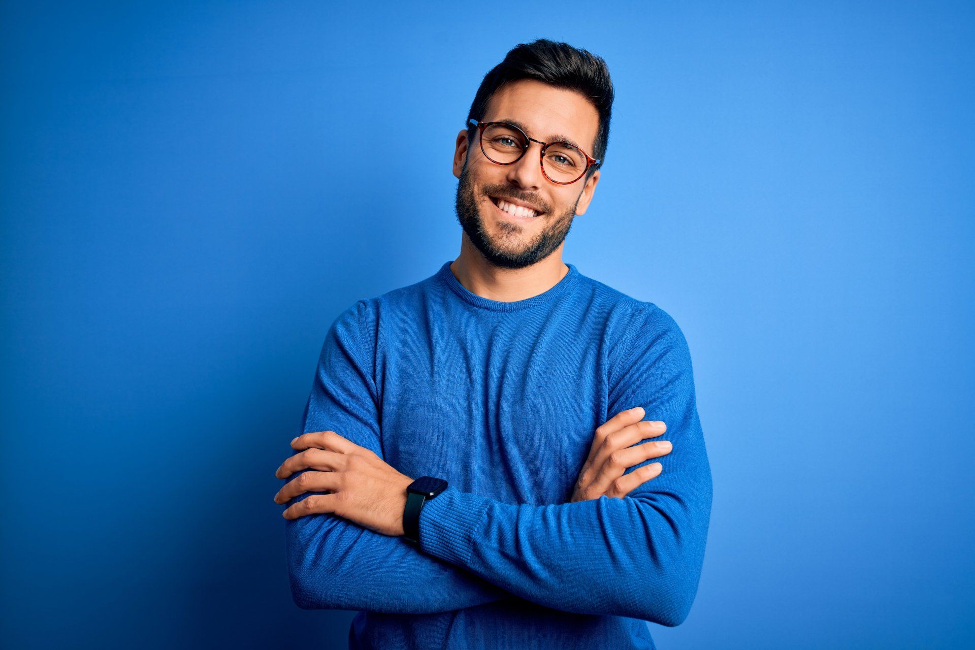 A person with a beard and glasses smiles, arms crossed, wearing a blue long-sleeved shirt against a solid blue background.