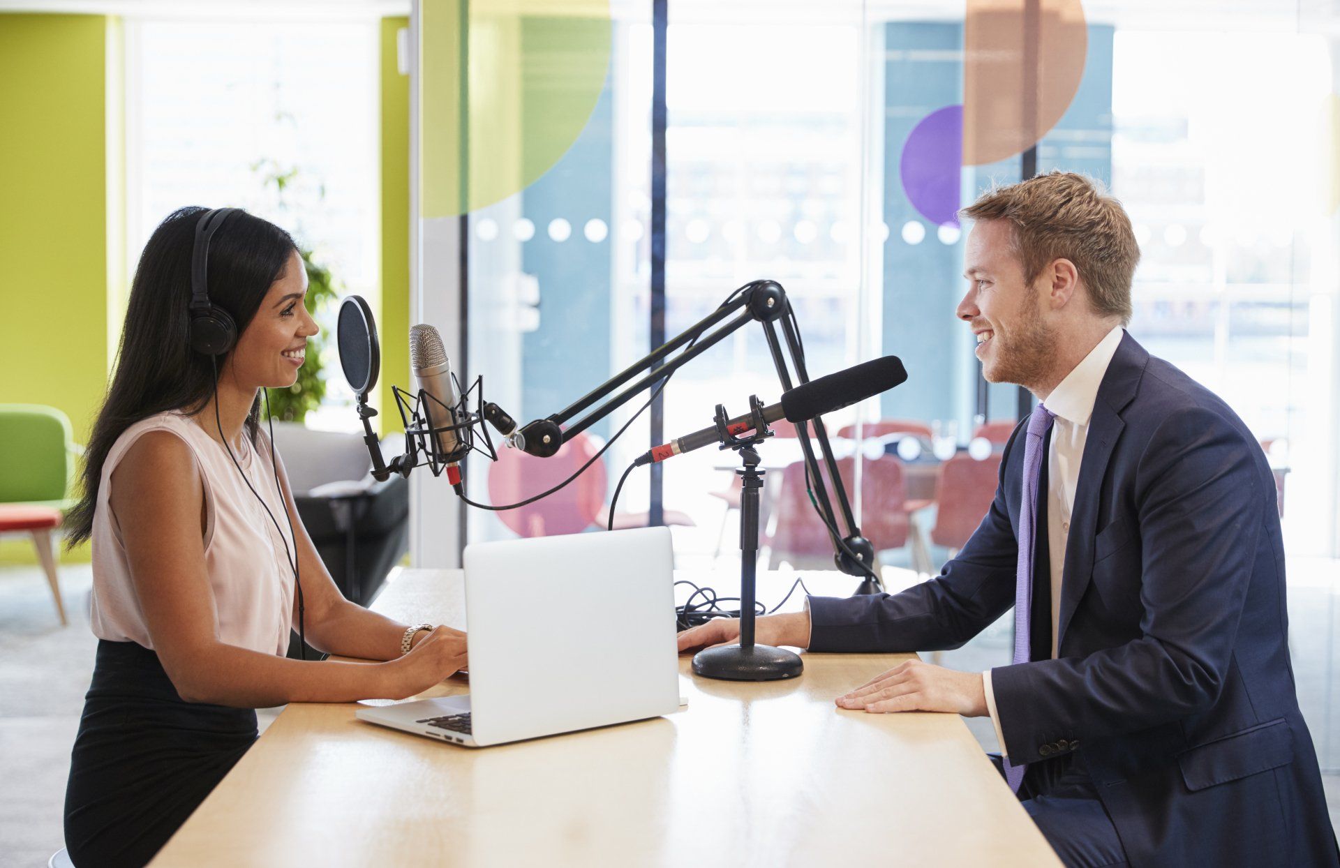 Two people sitting across from each other at a desk with microphones, recording a podcast in a modern office.