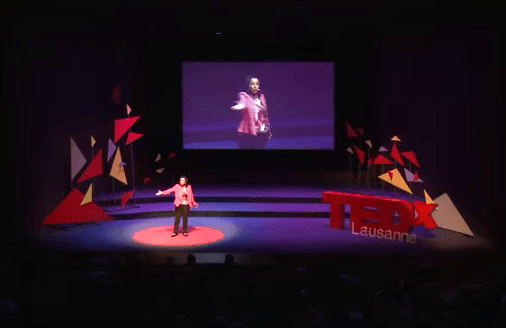 A speaker on a TEDx Lausanne stage gestures to a large screen displaying their image in a dimly lit auditorium.
