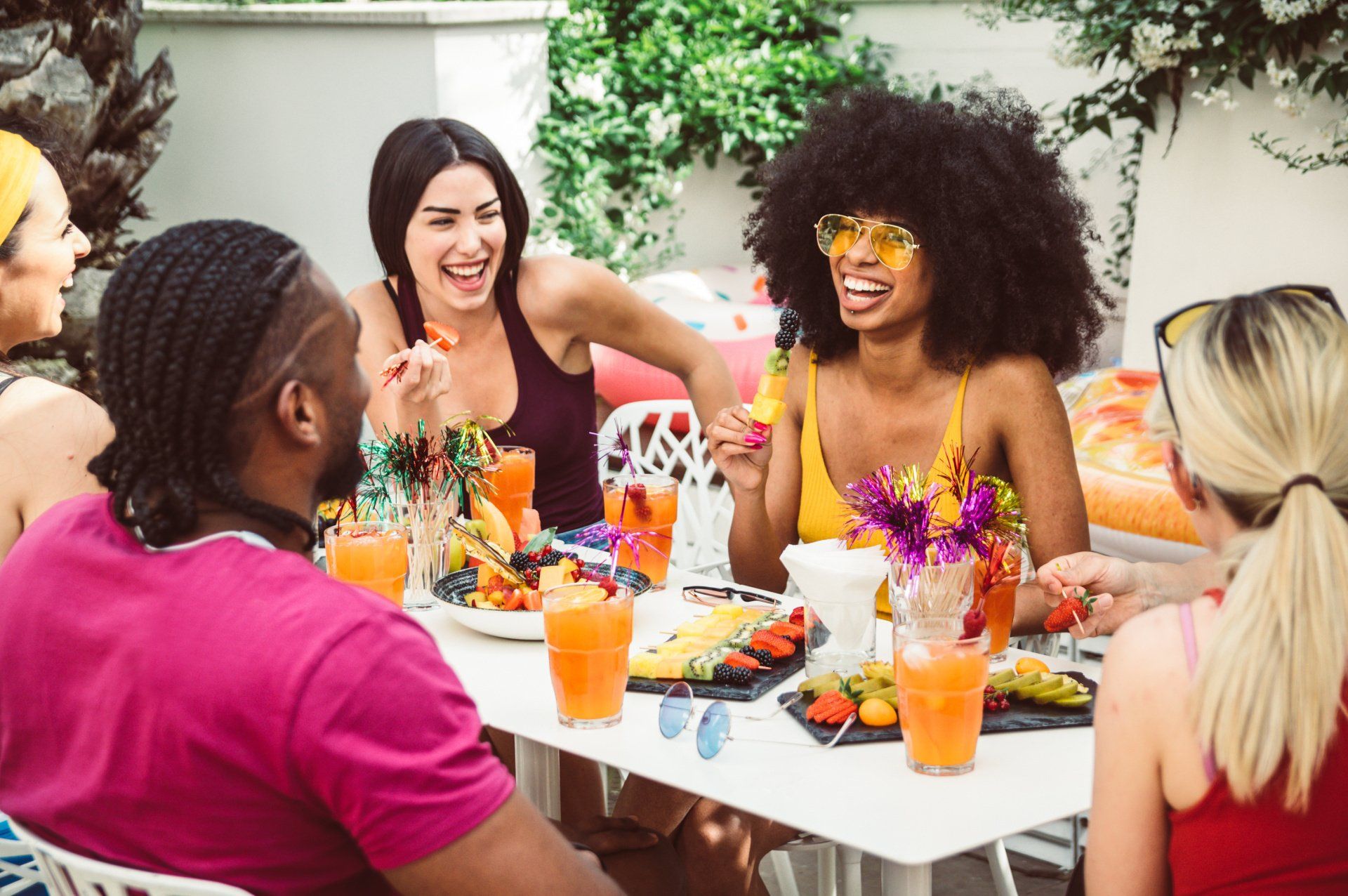 A photograph of a group of people, sat outside at a table with food and drinks, socialising and laughing with each other
