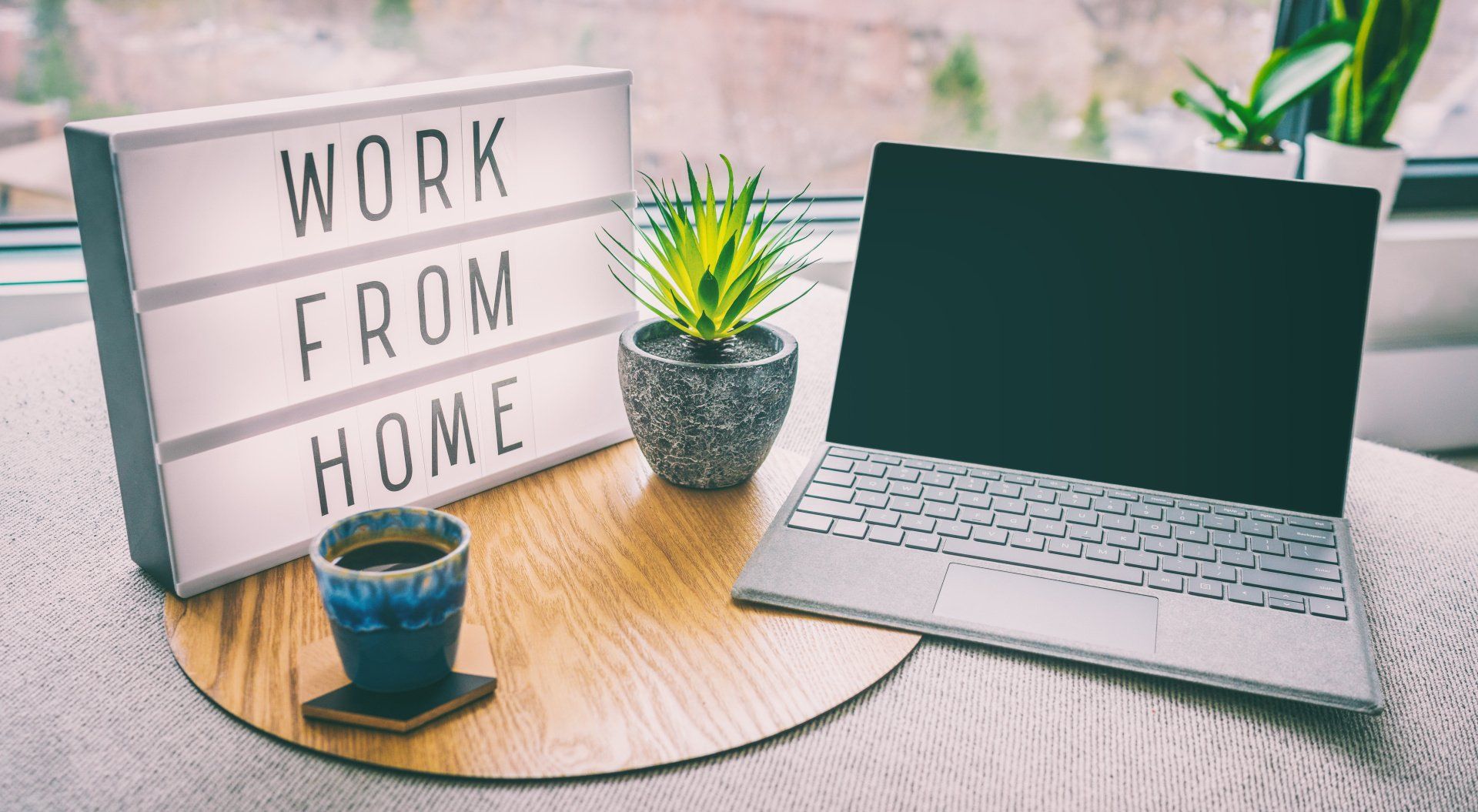 A small illuminated sign, that reads Work from Home, on a table next to an open laptop and cup of coffee