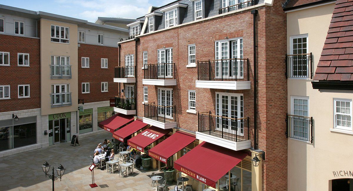 Red brick building with balconies, awnings, and a cafe. Beige and tan buildings in the background.