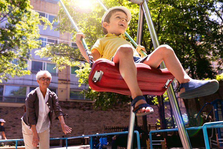 A young boy is sitting on a red swing in a park while an older woman watches.
