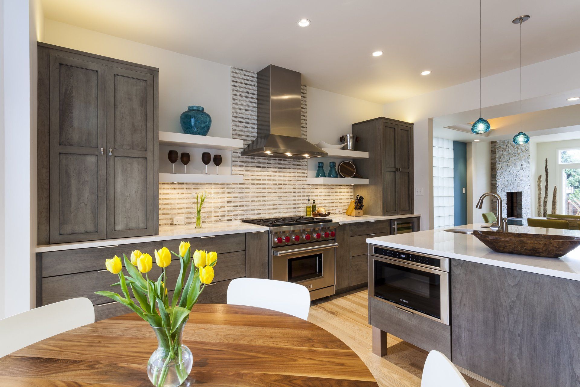 Modern kitchen with gray cabinets, white tile backsplash, stainless steel range, and a wooden dining table with yellow tulips