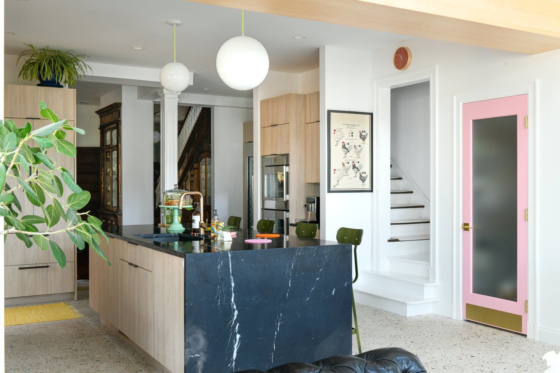 Modern kitchen with a black marble island, wood cabinetry, hanging globe lights, and a pink-framed door