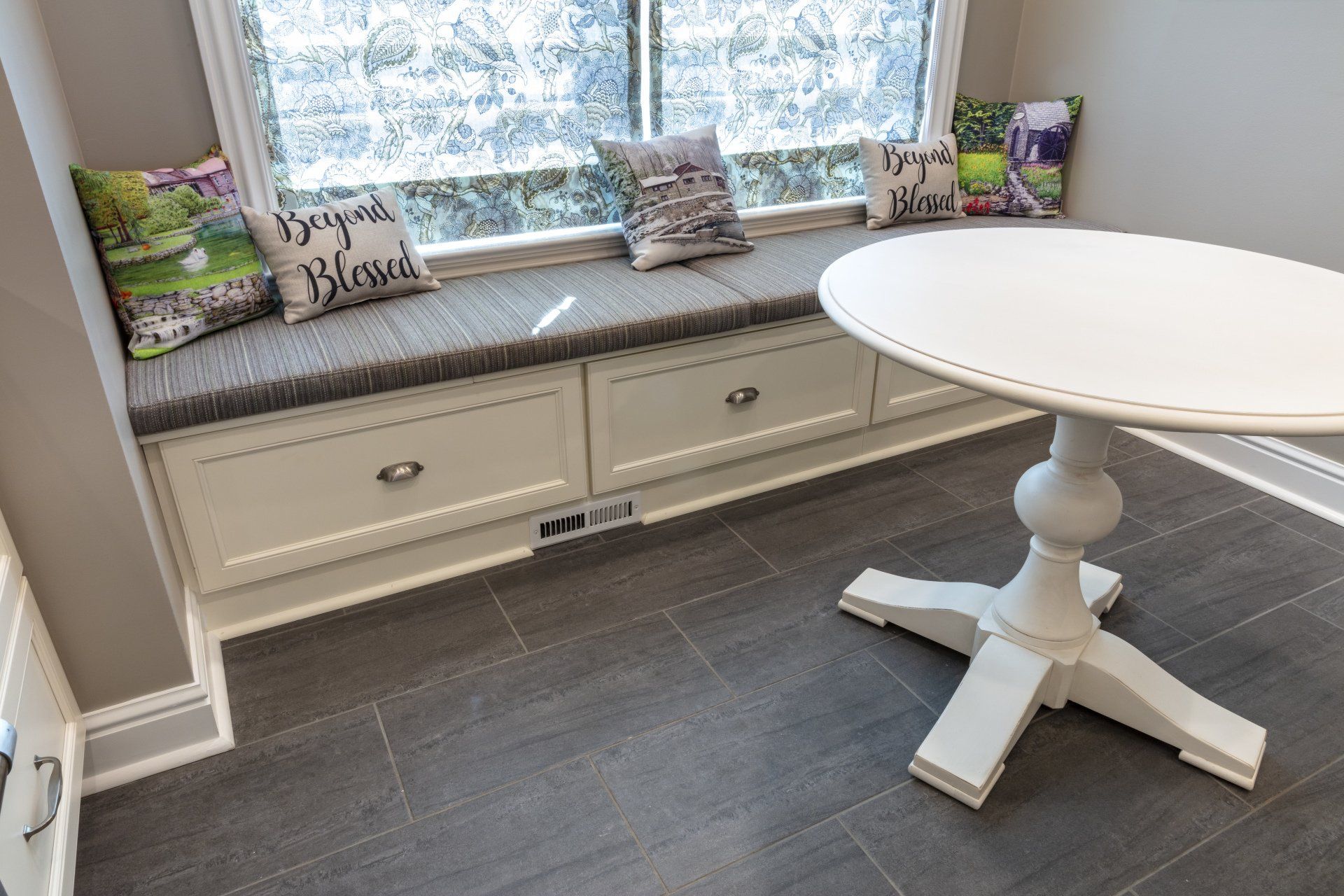 Window seat with gray cushions, decorative pillows, and a round white table on dark tile flooring.
