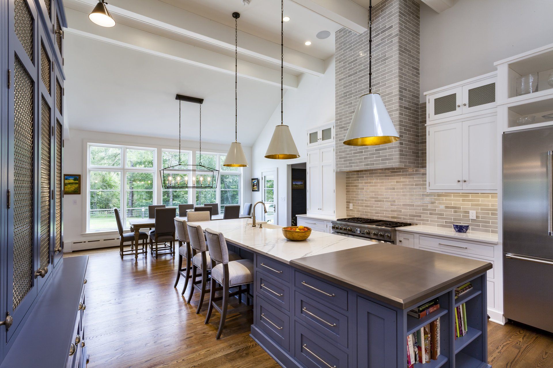 Modern kitchen with dark blue island, pendant lights, brick backsplash, and dining area by large windows.