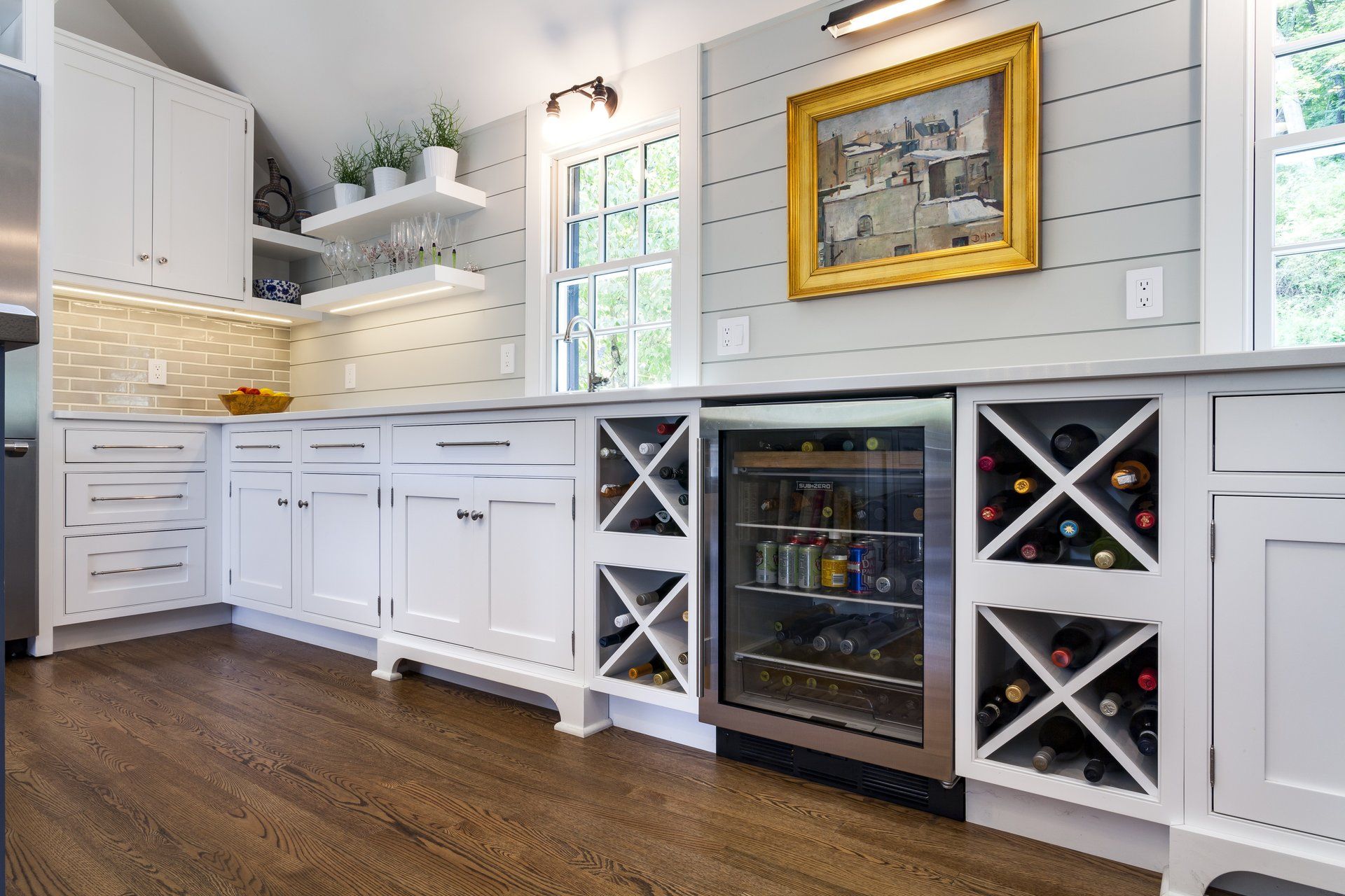 Bright white kitchen with wine fridge, X-shaped bottle racks, dark wood floor, and gold-framed wall art