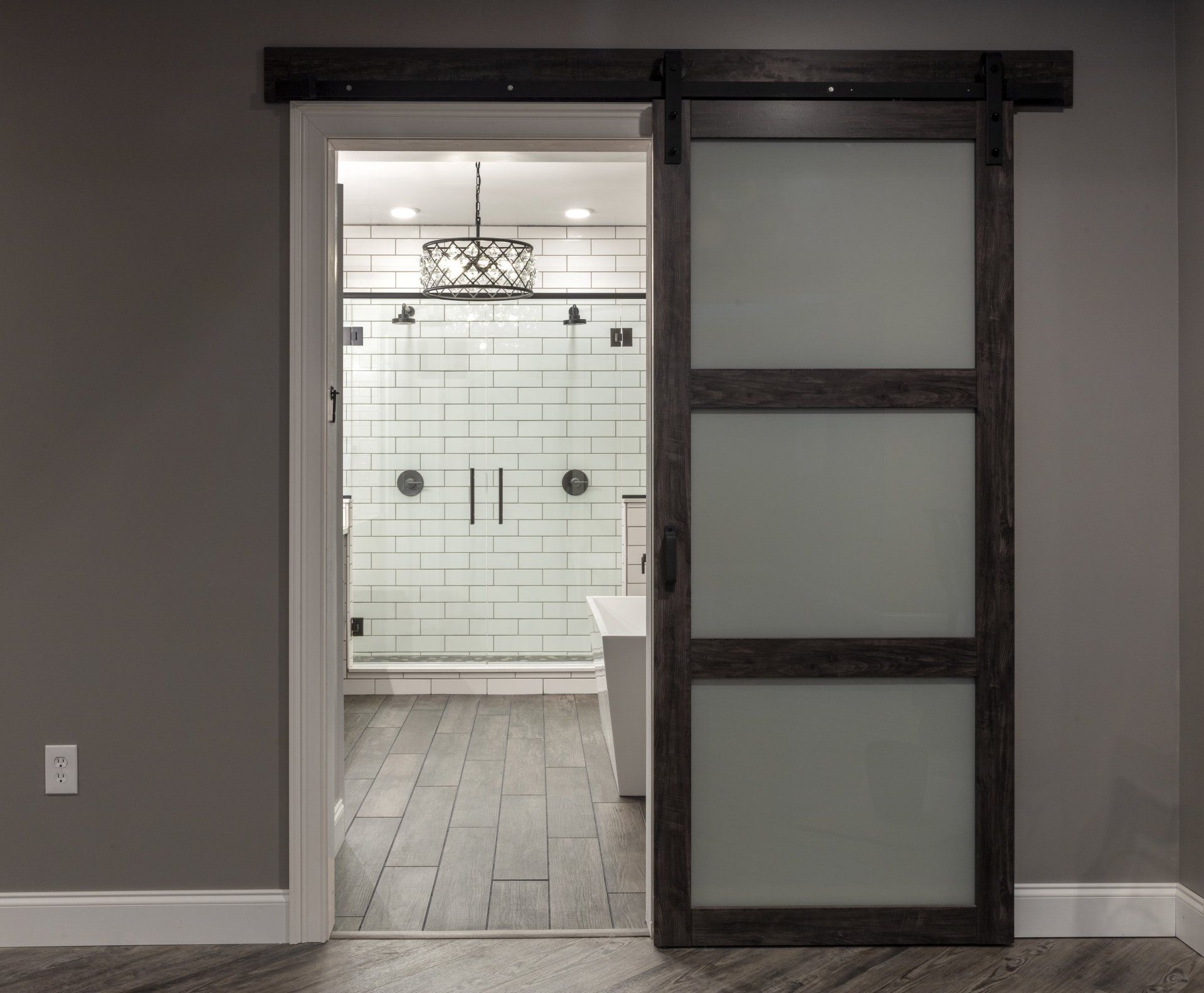 Barn-style sliding door opening to a bright tiled bathroom with glass shower and chandelier.
