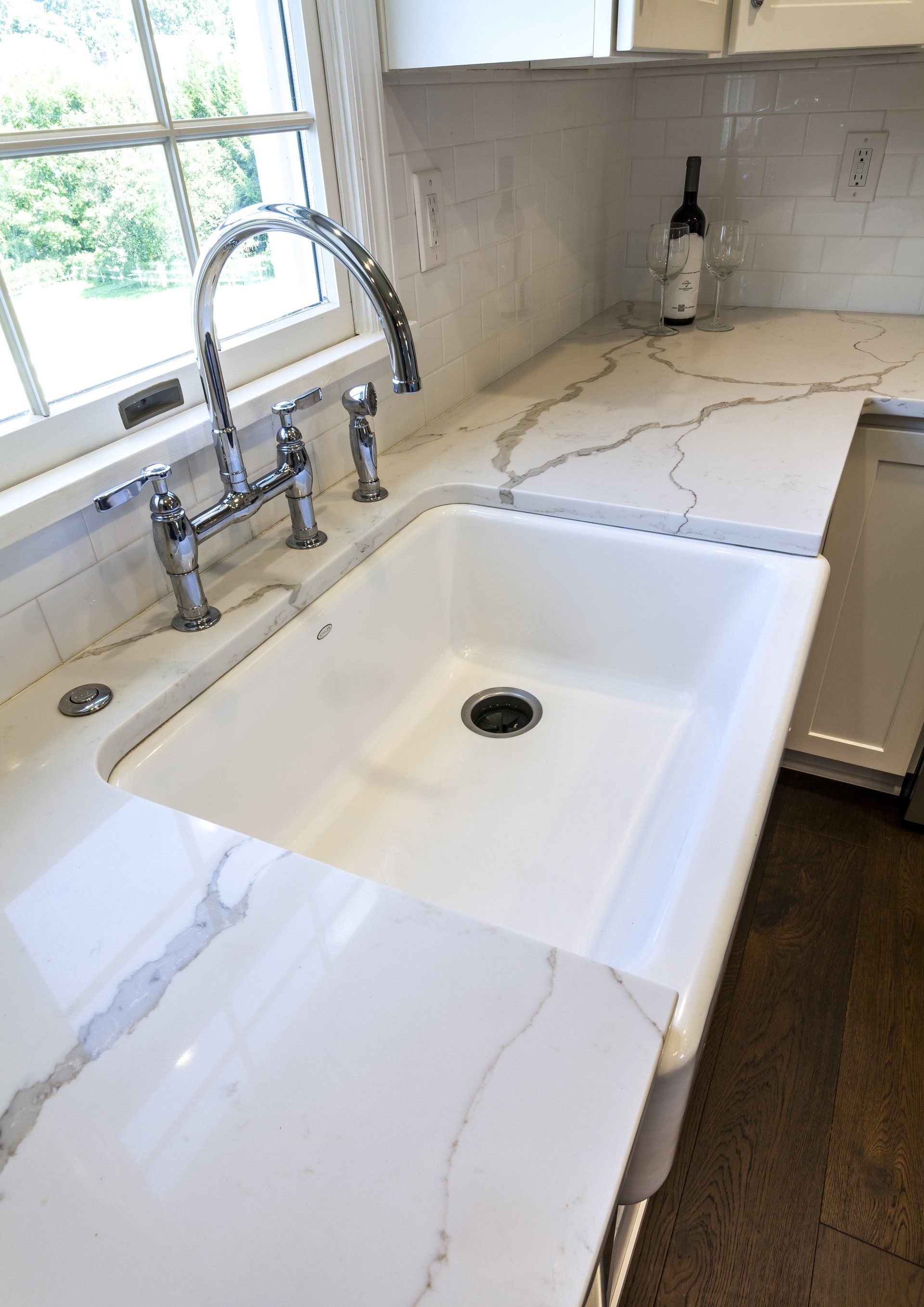 White farmhouse kitchen sink with chrome faucet and marble countertop beside a window