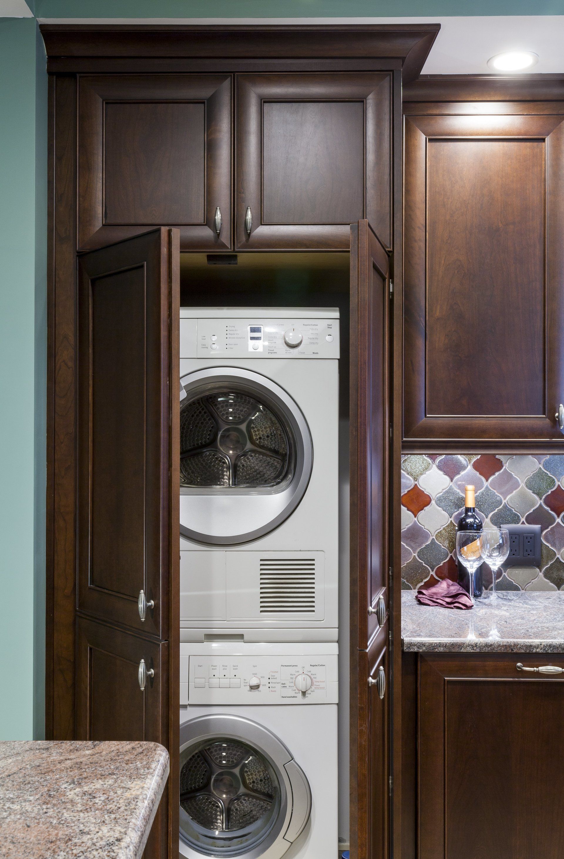 Stacked washer and dryer in a dark wood laundry cabinet, partly open in a kitchen.