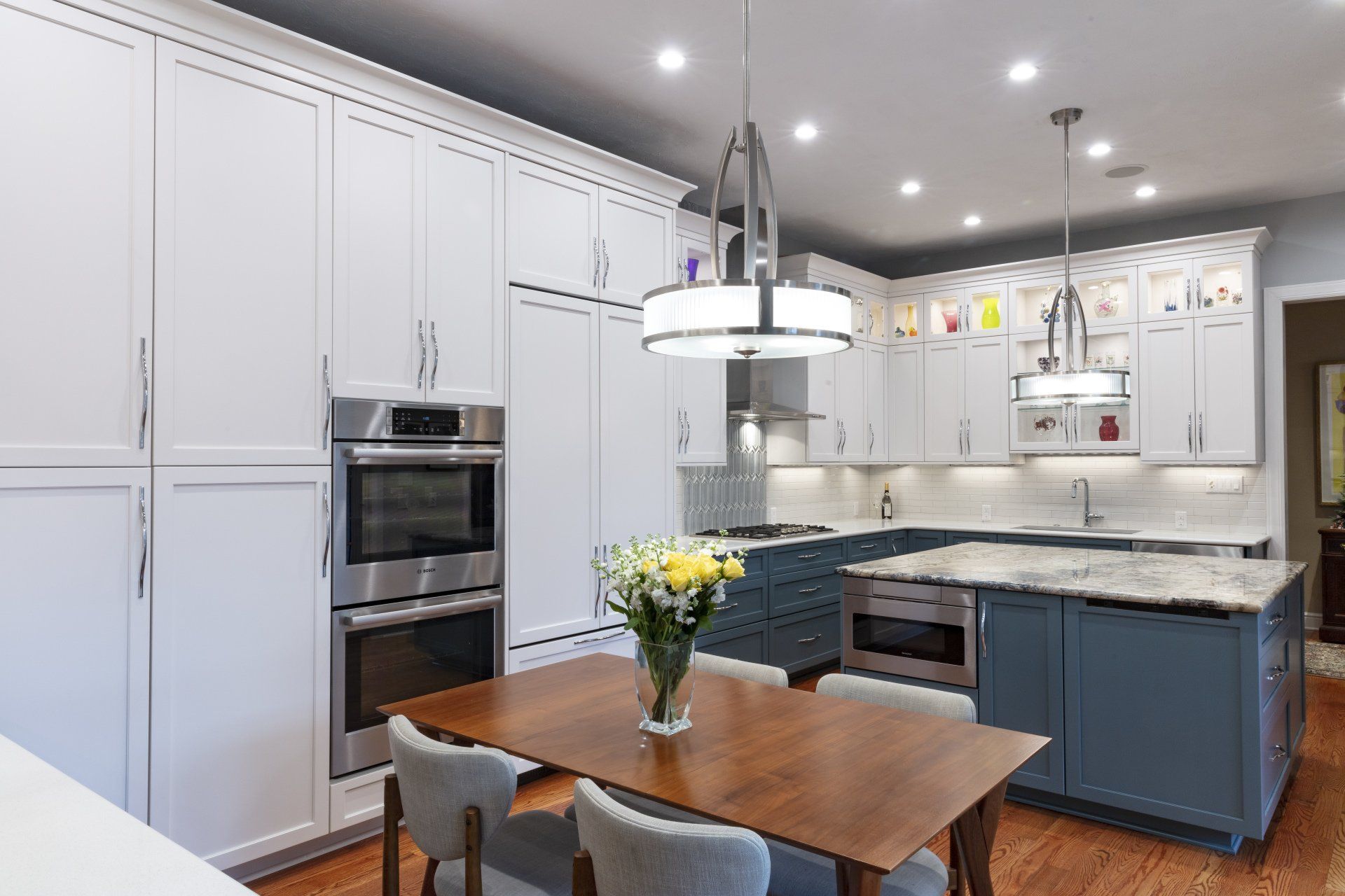 Modern white kitchen with blue island, marble countertops, pendant light, and wooden dining table with flowers