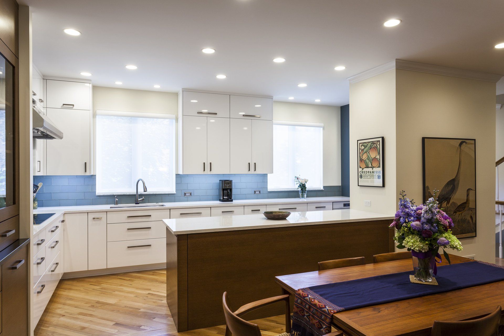 Bright modern kitchen with white cabinets, blue backsplash, and a wooden dining table with flowers.