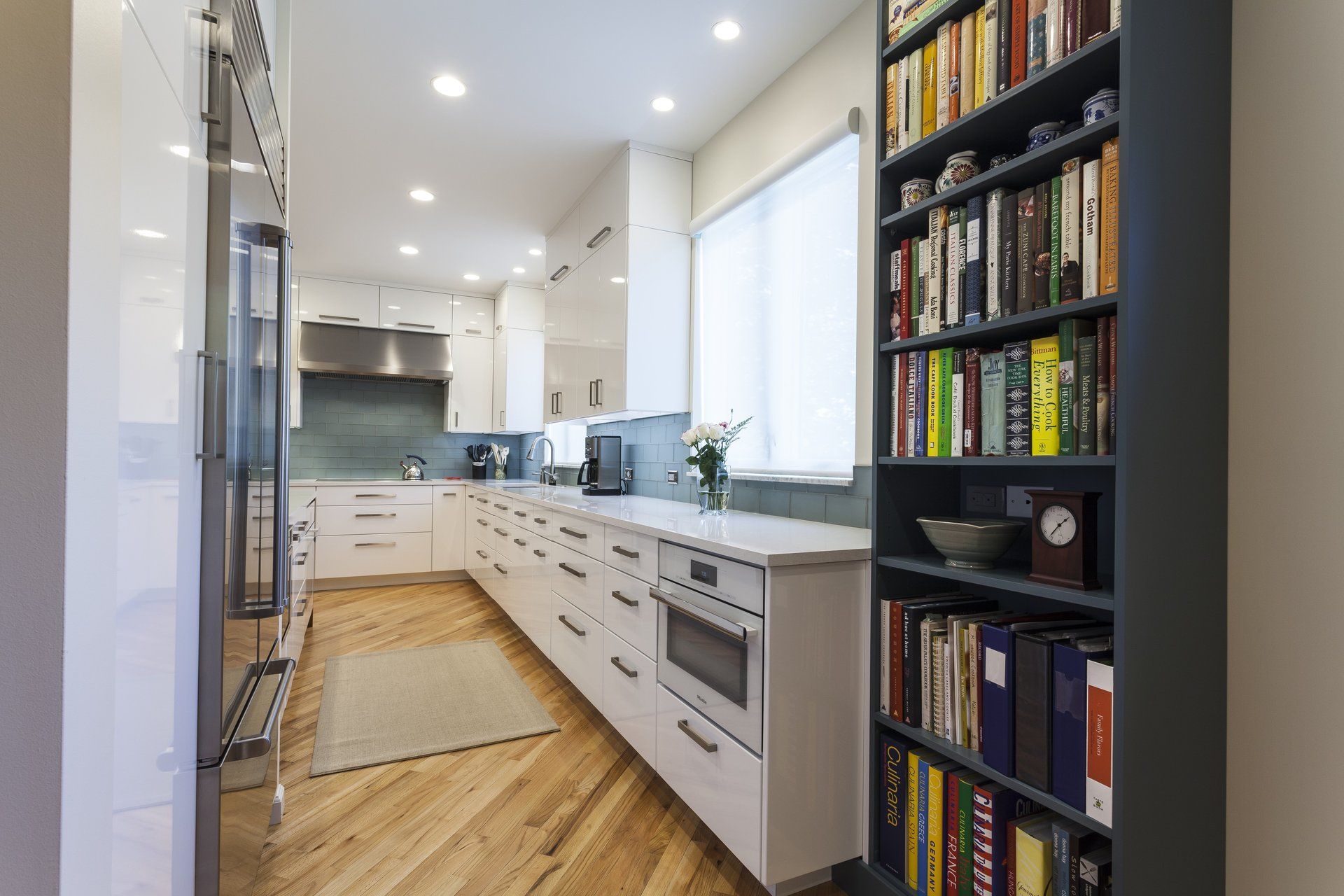 Bright modern galley kitchen with white cabinets, hardwood floor, stainless appliances, and a bookshelf nook