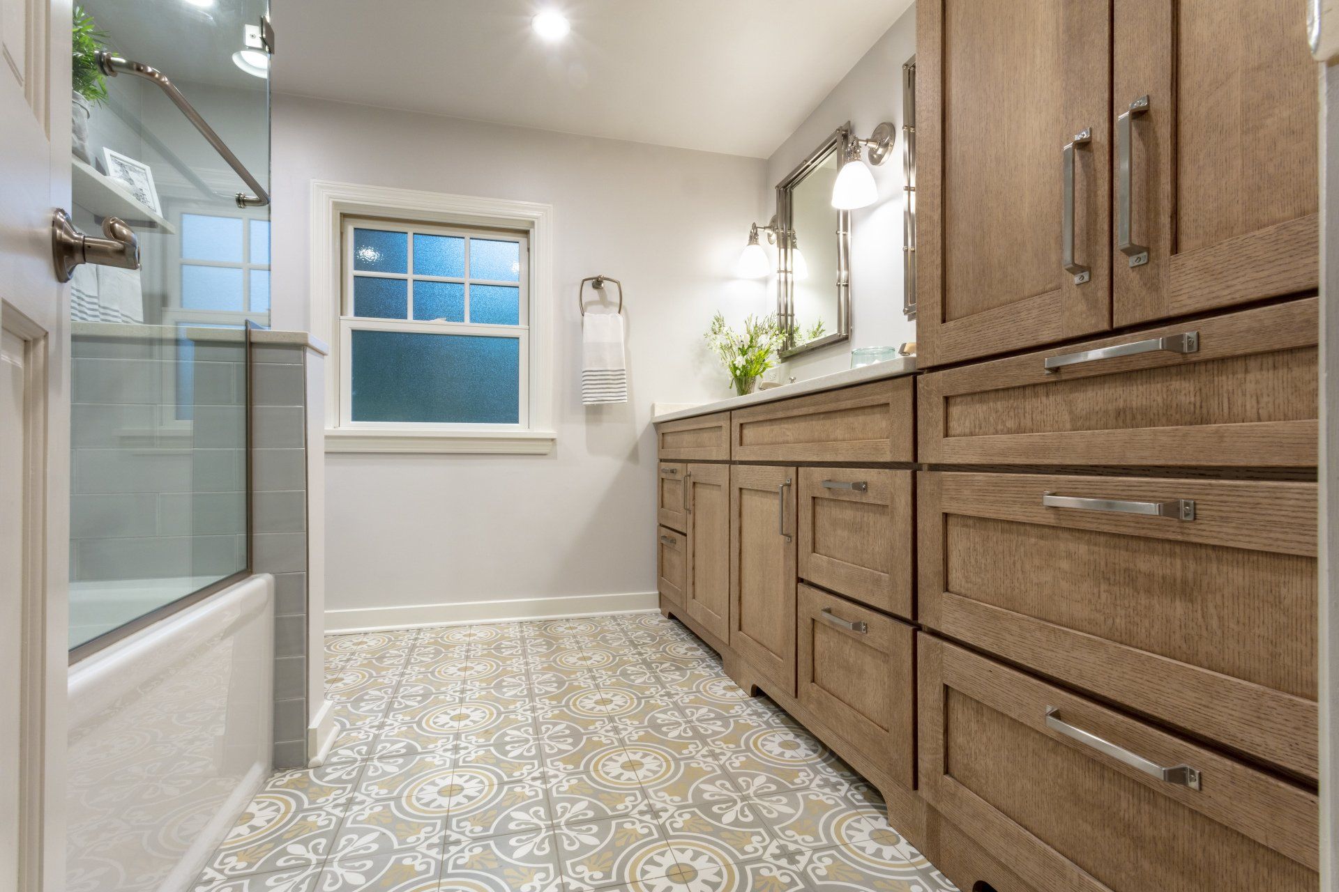 Bright bathroom with wooden vanity, patterned tile floor, shower, and a window with a plant.