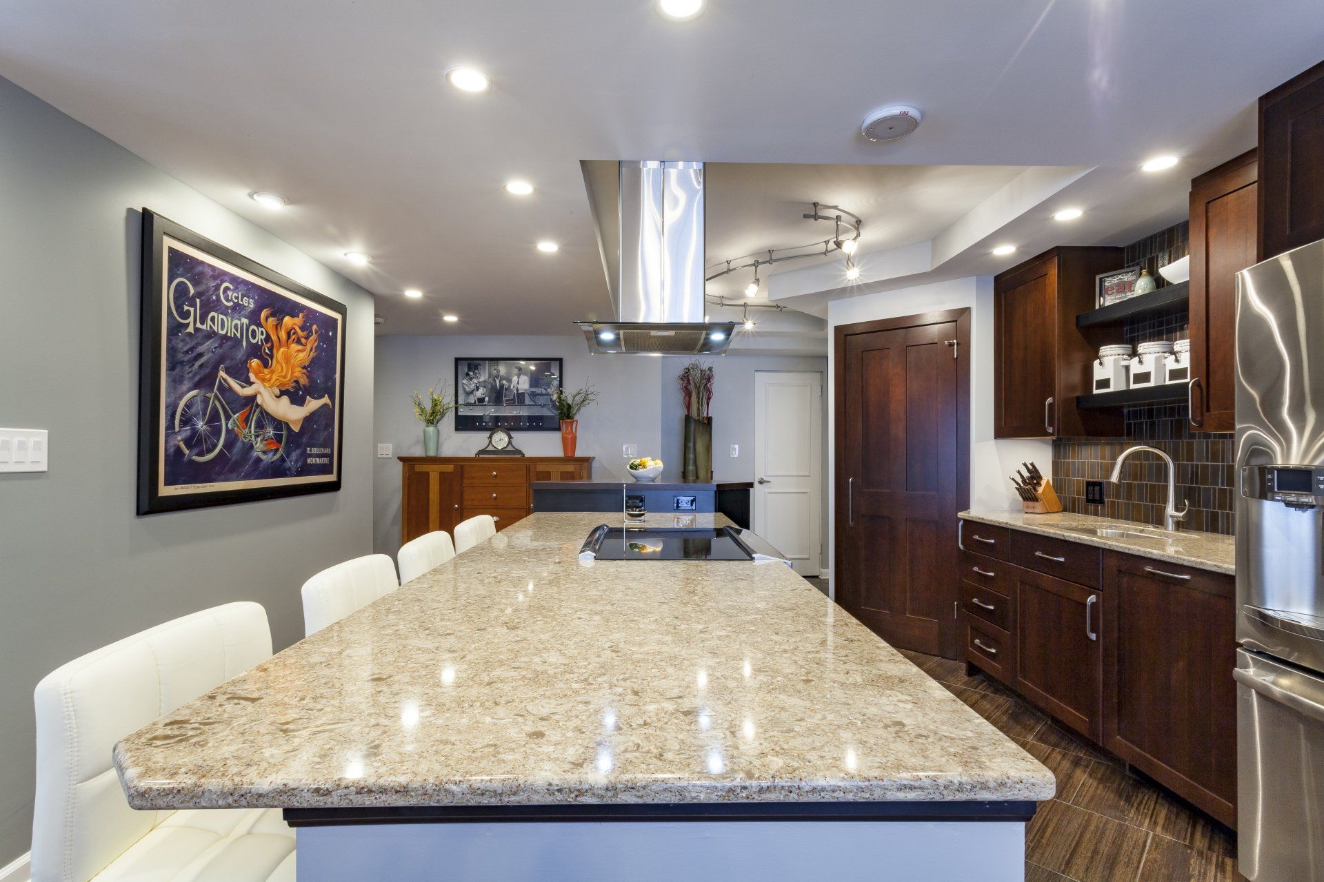 Modern kitchen with a granite island, white chairs, dark wood cabinets, and recessed ceiling lights