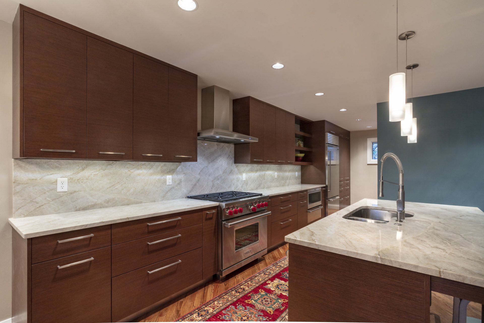 Modern kitchen with dark wood cabinets, marble counters, stainless stove, and island sink under pendant lights