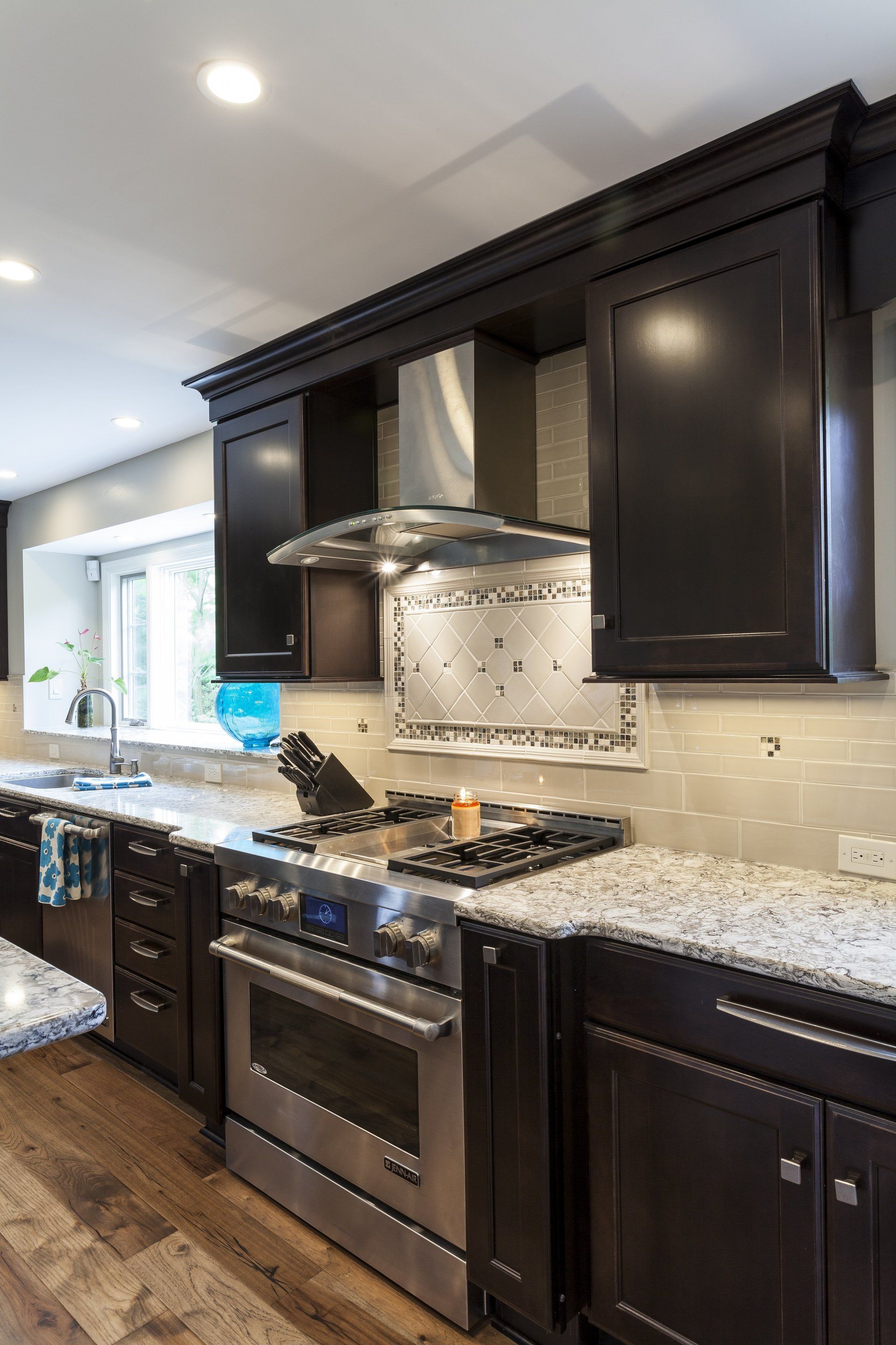 Modern kitchen with dark cabinets, stainless appliances, granite countertops, and a stove under a metal hood.