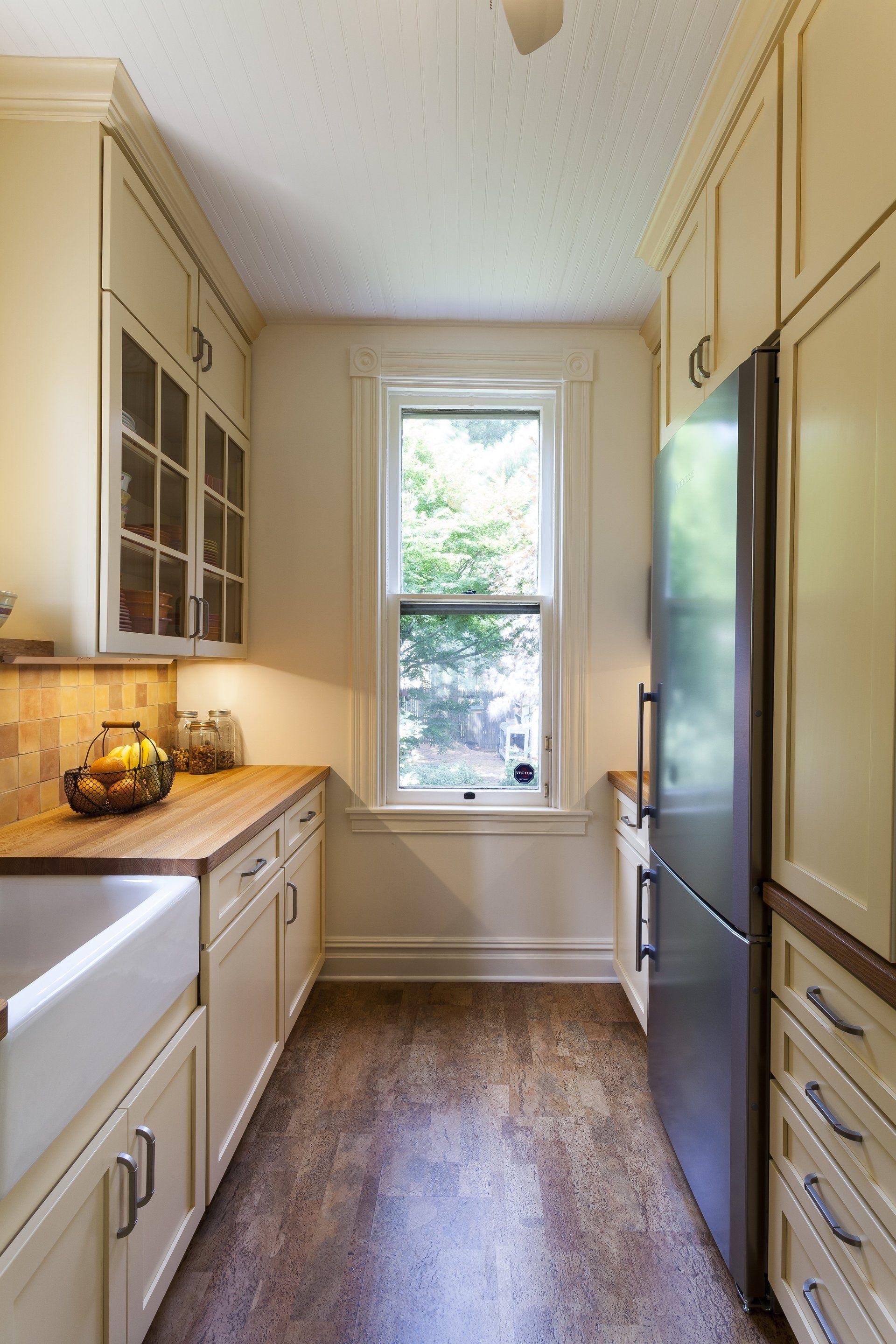 Narrow galley kitchen with white cabinets, wood floors, black refrigerator, and a window at the end.