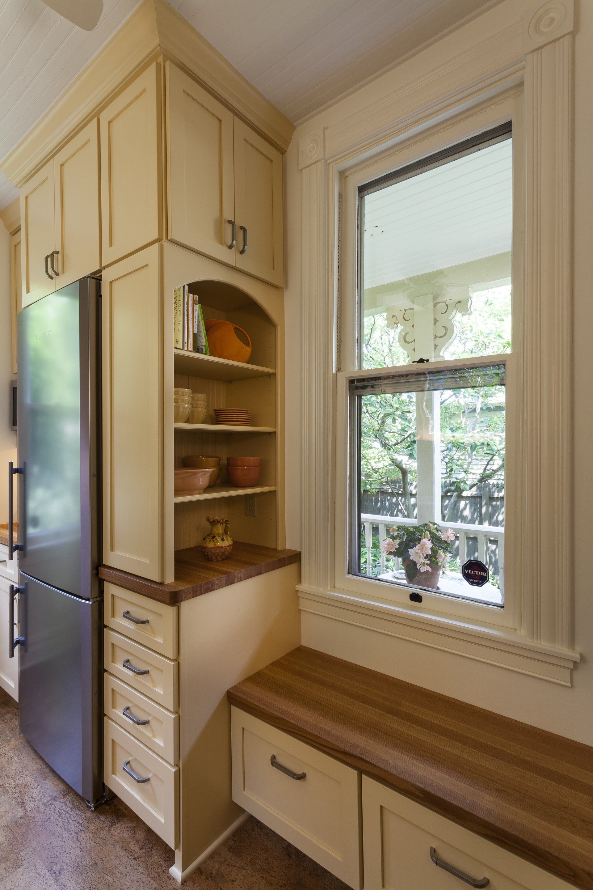 Narrow kitchen nook with built-in pantry shelves, drawers, and a window seat beside a refrigerator.