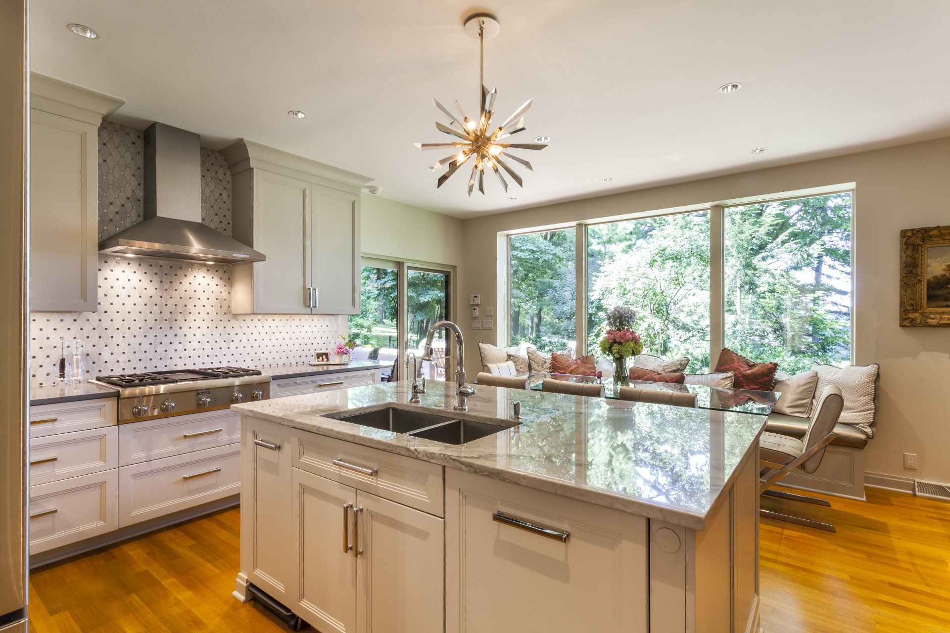 Bright modern kitchen with large island, white cabinets, stainless range hood, and big windows overlooking trees