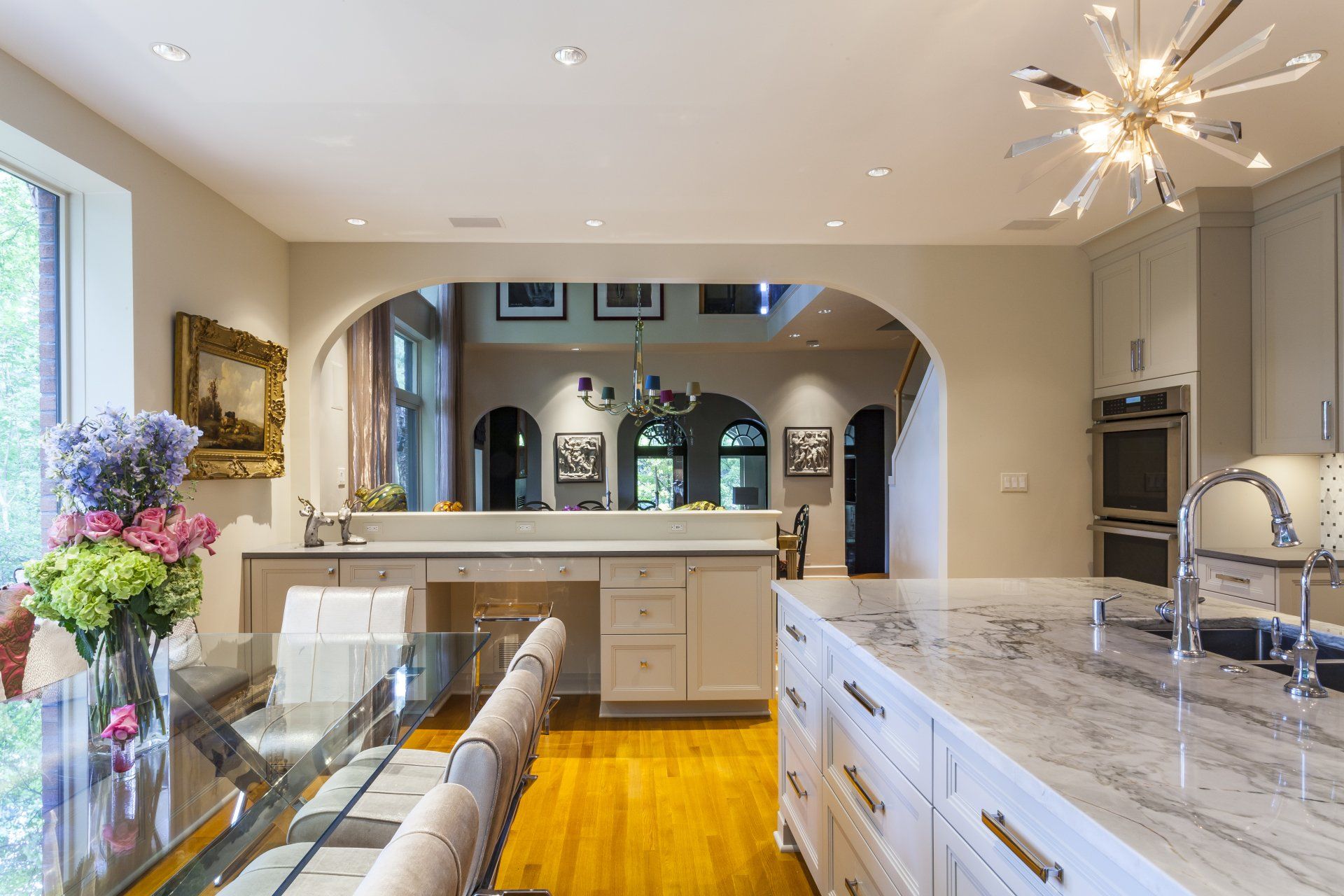 Bright modern kitchen with marble island, white cabinets, flowers, and hardwood floors opening to an arched living area
