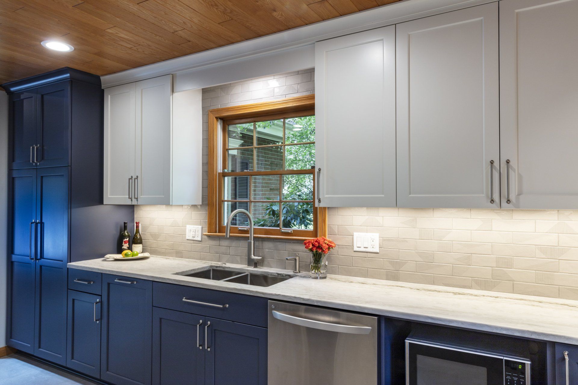 Modern kitchen with blue and gray cabinets, window above sink, and wood-paneled ceiling
