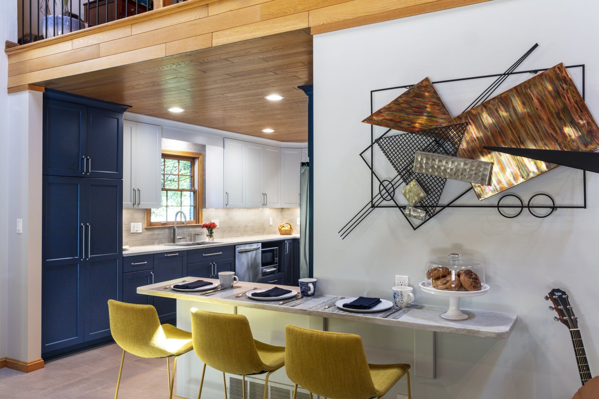 Modern kitchen and dining nook with blue cabinets, white table, yellow chairs, and wall art above the table
