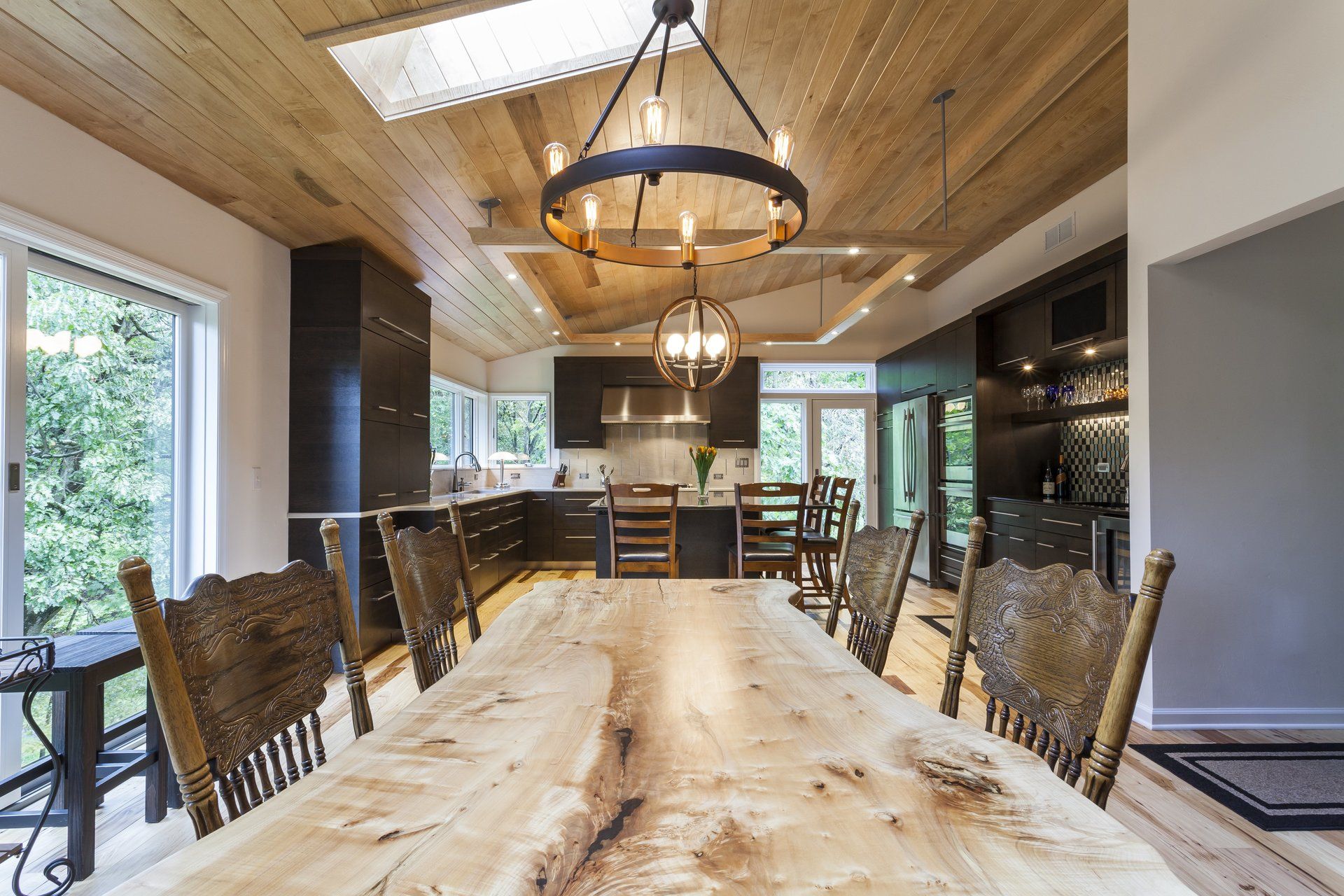 Modern dining room with a long wood table, rustic chairs, and large windows under a wooden ceiling