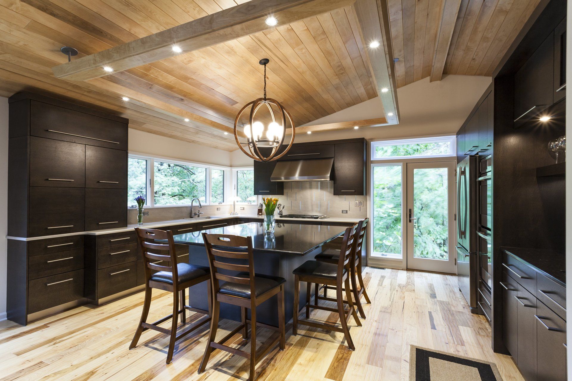 Modern kitchen with dark cabinets, wood ceiling, island seating, and large windows overlooking greenery