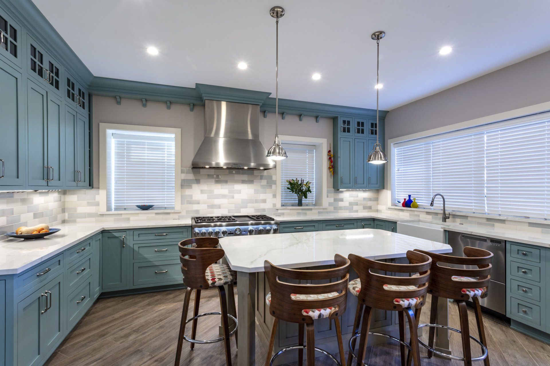 Bright kitchen with blue cabinets, white island, stainless range hood, and bar stools by large windows.